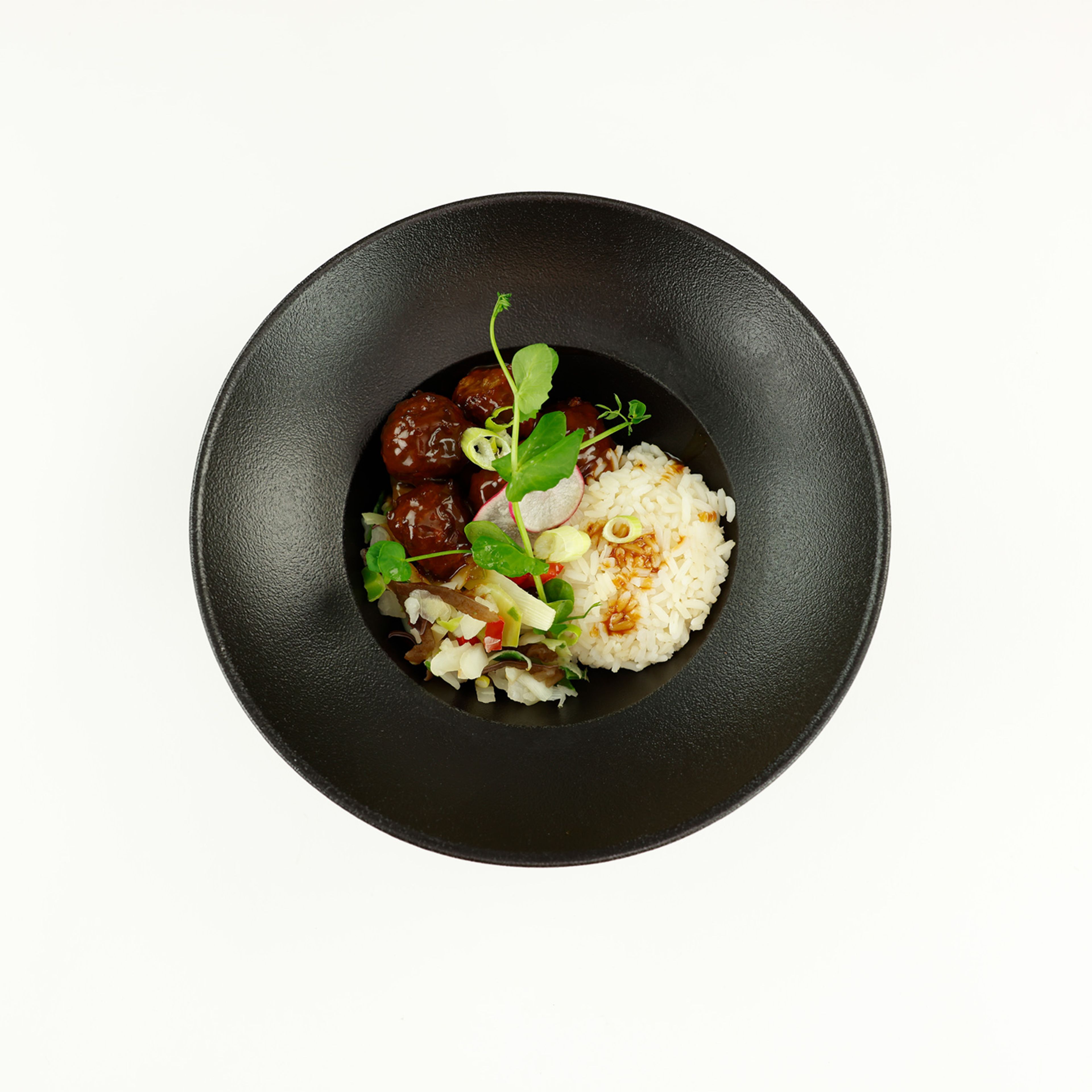 Photograph of a deep black bowl featuring a culinary presentation of rice, garnished with crispy shallots, and meatballs covered in sauce, served alongside a salad of fresh vegetables and edible leaves. The composition highlights the contrast between the light-colored ingredients and the dark bowl, emphasizing refined and modern food styling.