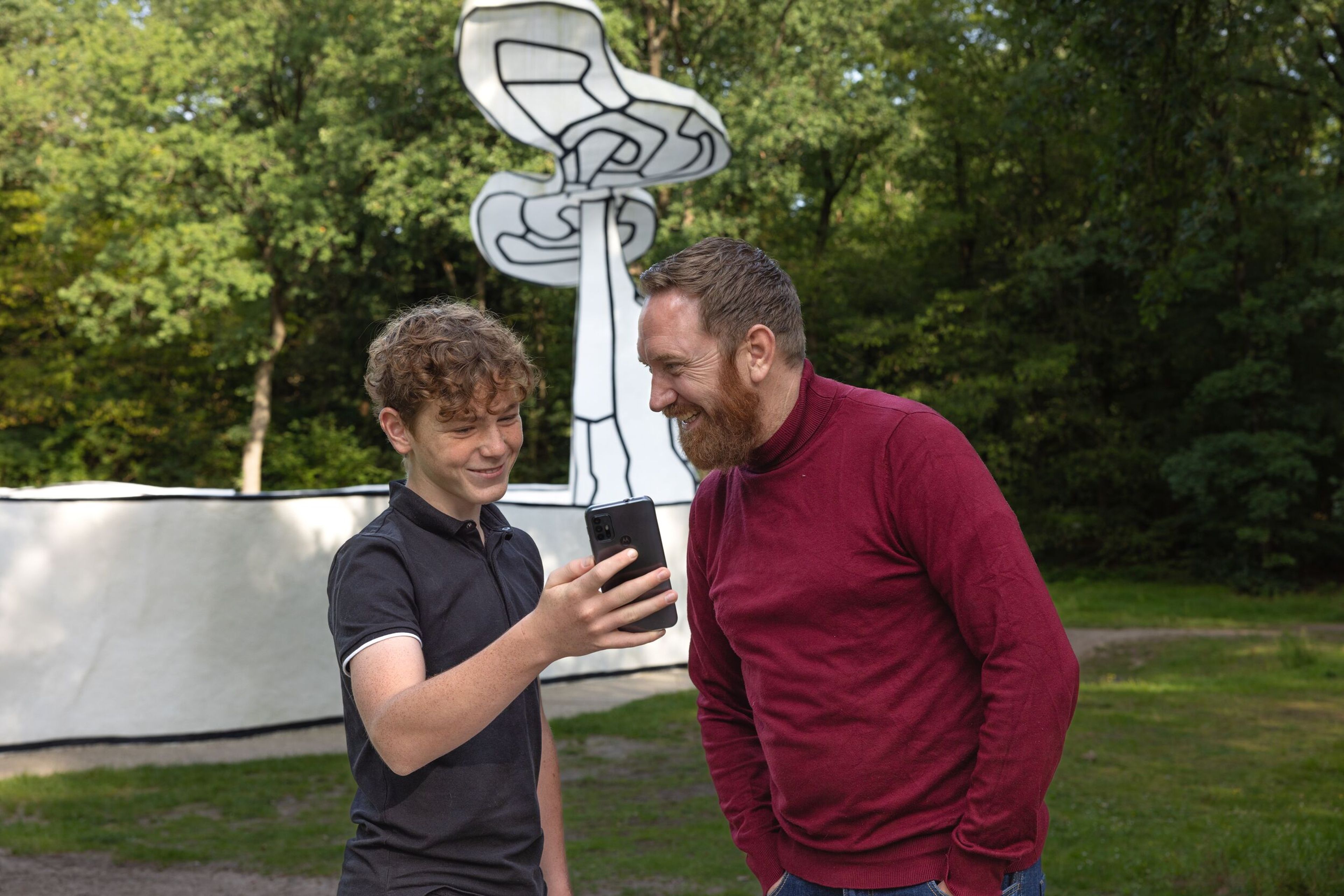 Kind en volwassene staan bij Jardin d'émail. Kind heeft mobiel in hand