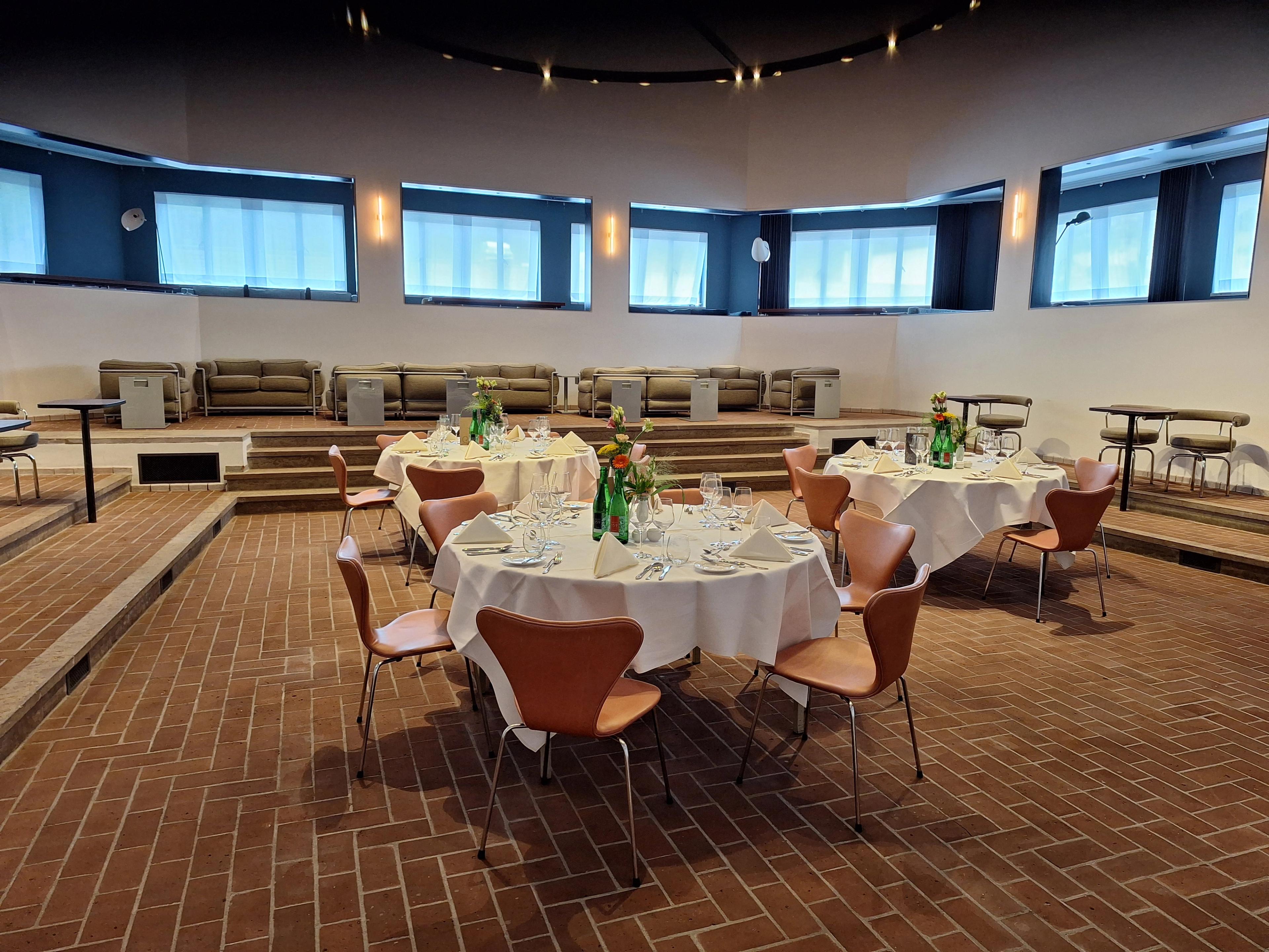 Foto van een aantal ronde gedekte tafels met stoelen in een halfronde ruimte met uitzicht naar buiten. Op tafel bloemen.