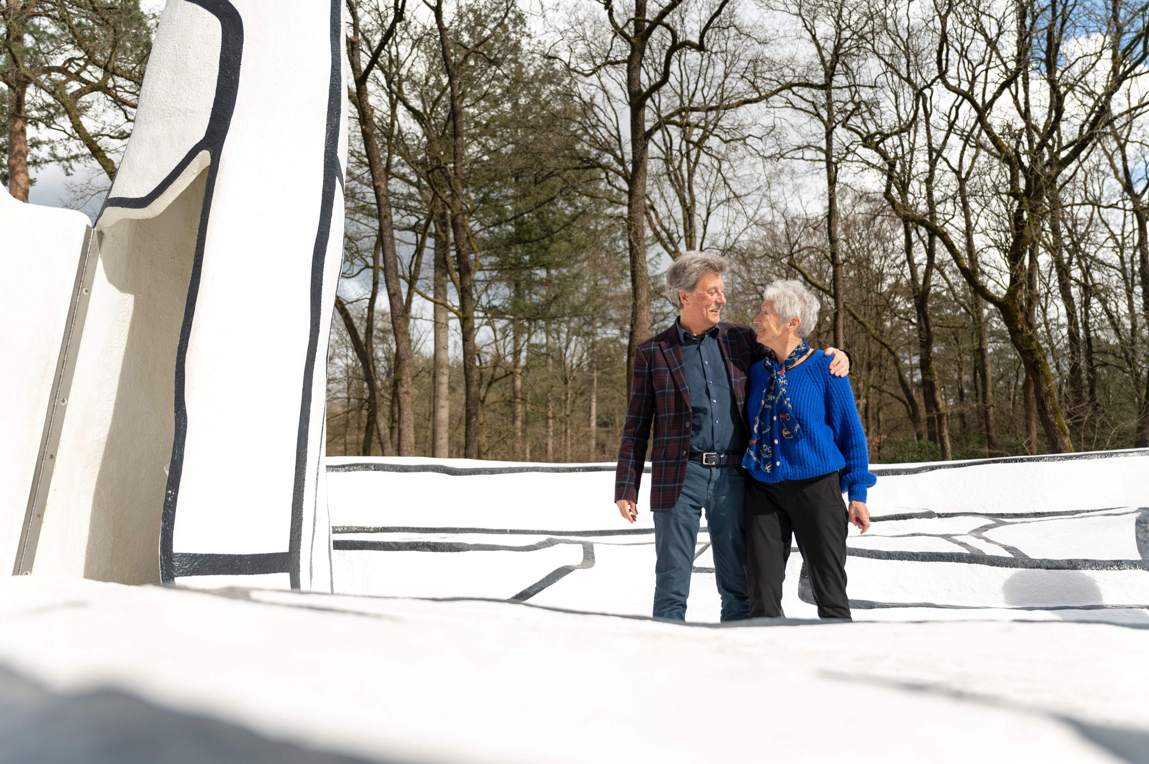 Foto van twee personen op een zwartwitte ondergrond, een kunstwerk. Op de achtergrond bomen. De man heeft een arm om de vrouw heen geslagen, ze lachen naar elkaar.