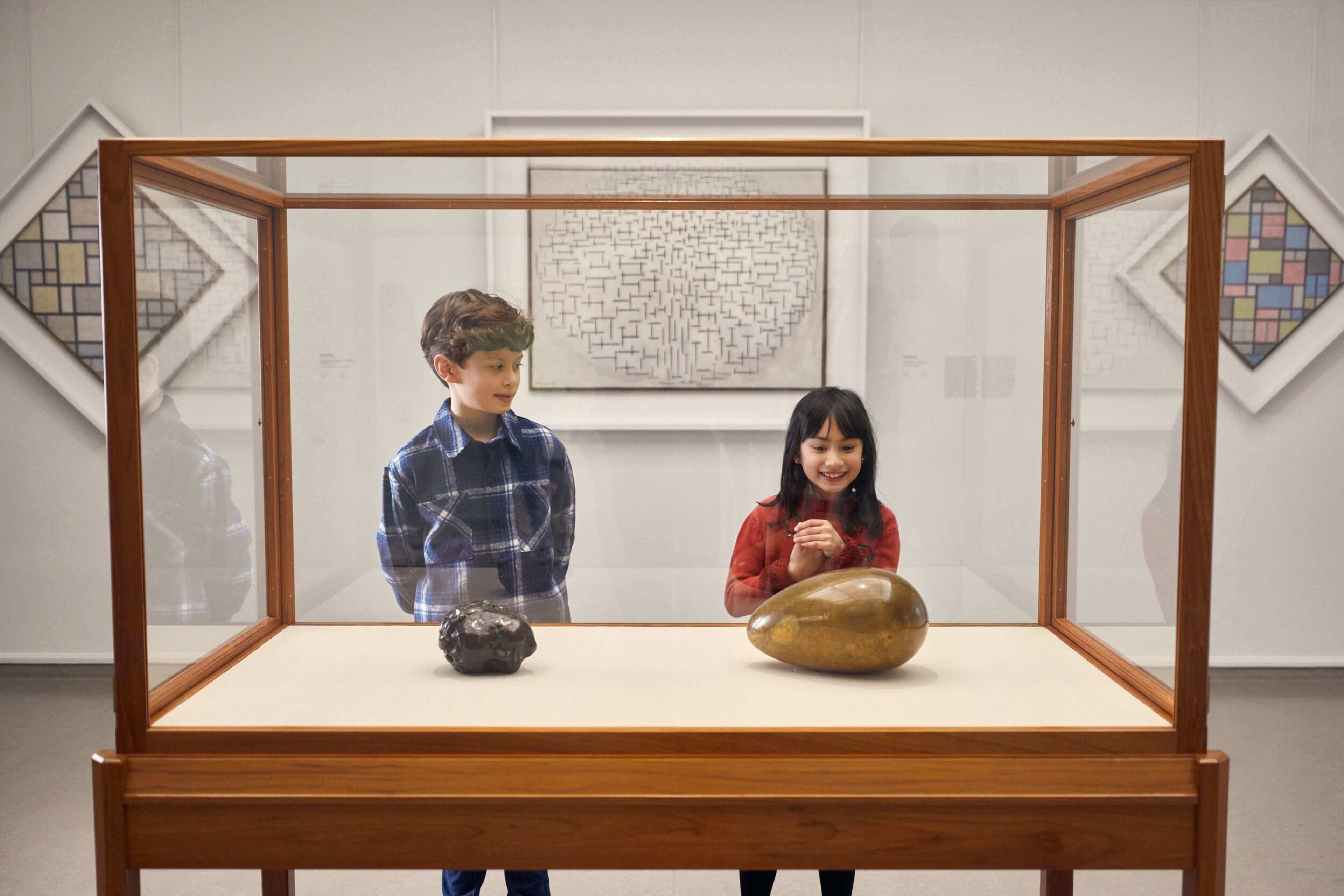 Foto van twee kinderen die een houten vitrinekast in een museum bekijken met daarin twee sculpturen, een donker hoofdje en een goudkleurig ei. Op de achtergrond hangen abstracte kunstwerken met geometrische patronen in verschillende kleuren en vormen.