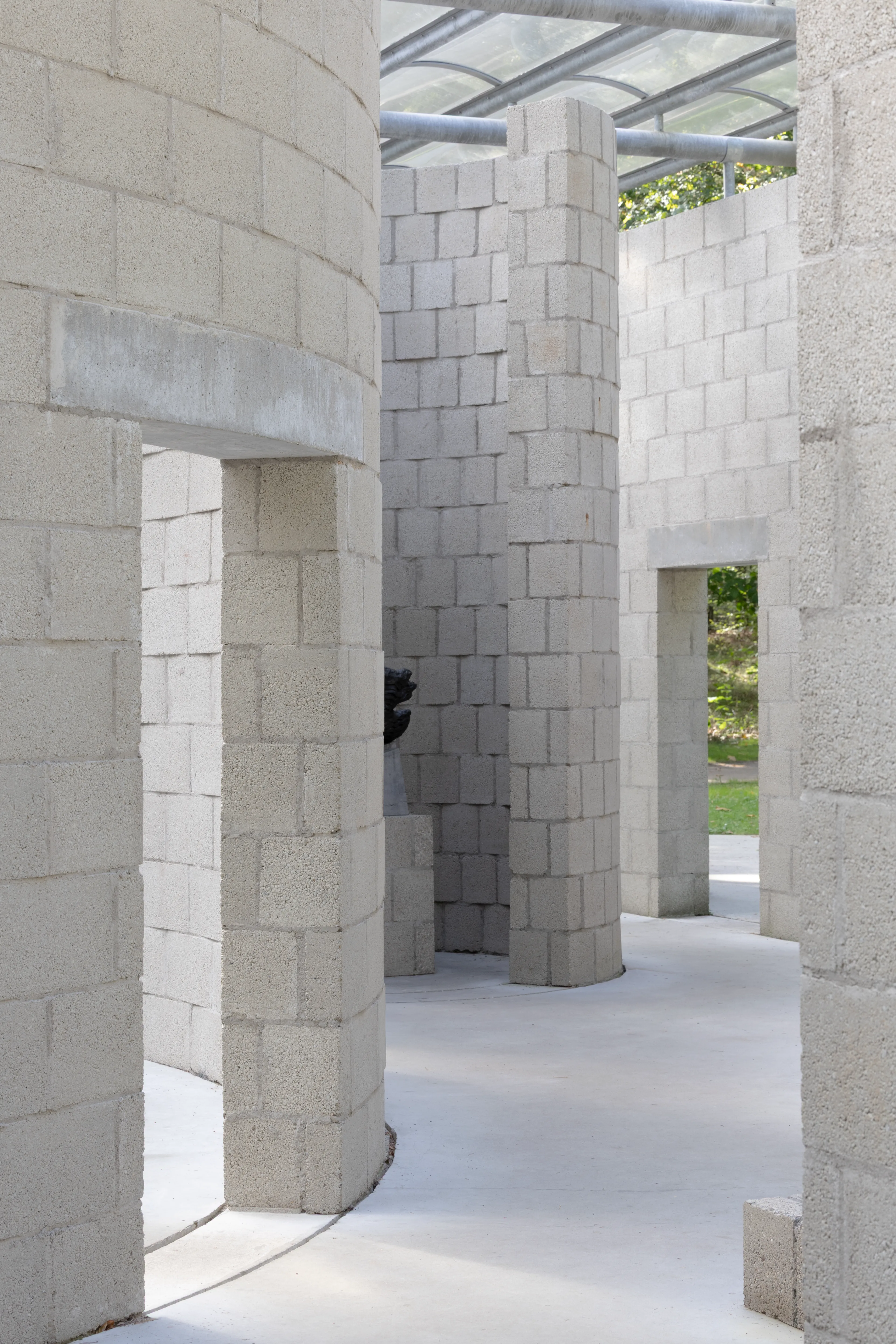 Photo of a modern exterior wall structure made of concrete blocks, featuring a transparent, undulating canopy of glass or plastic. Shadows from the surrounding trees fall on the walls