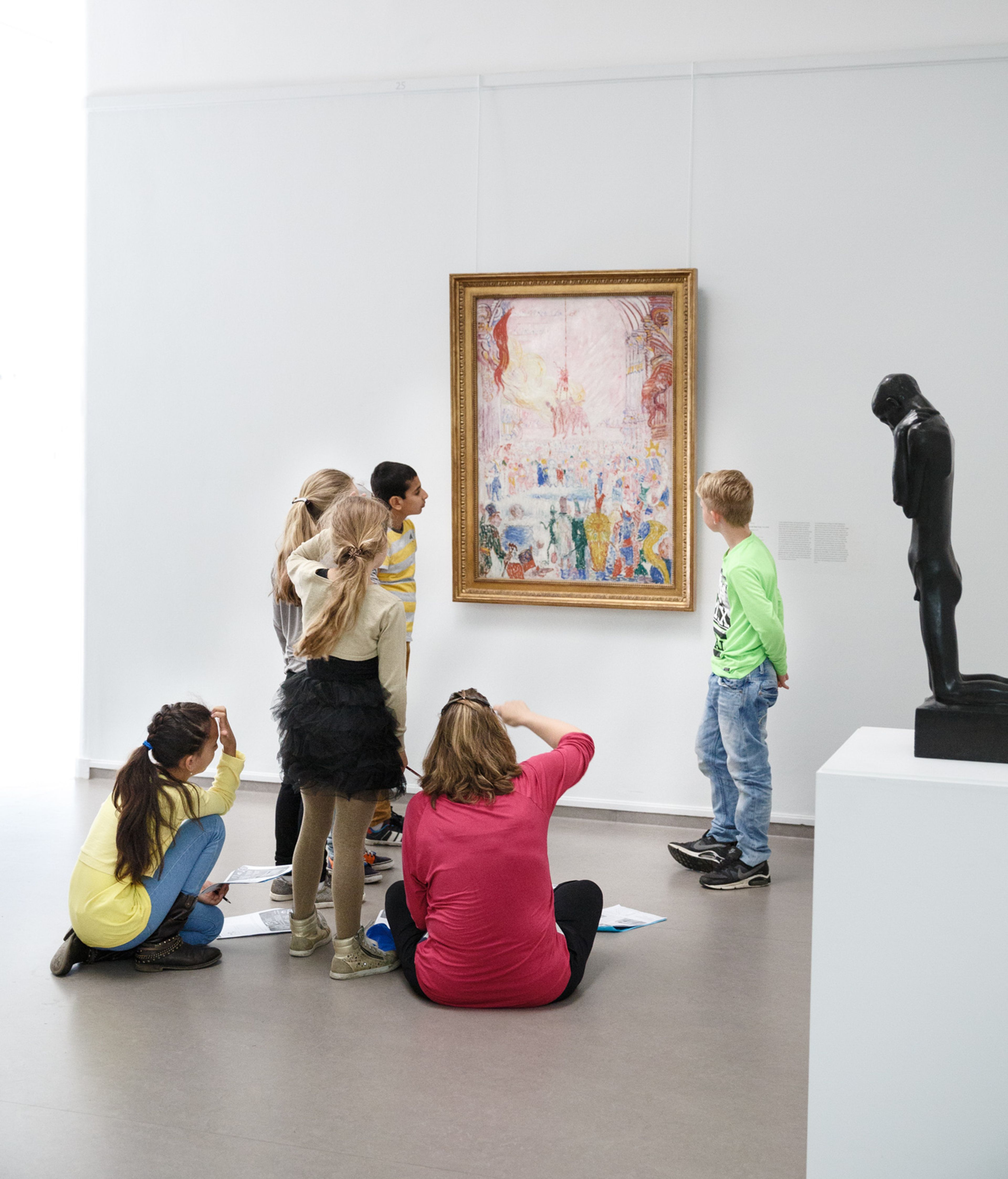 Groep kinderen bekijkt en bestudeert een schilderij in een museumruimte met witte muren en een grijze vloer. Links zitten drie kinderen op de grond, rechts staan drie kinderen, terwijl een zwarte beeldhouwwerk op een sokkel rechts in beeld staat.