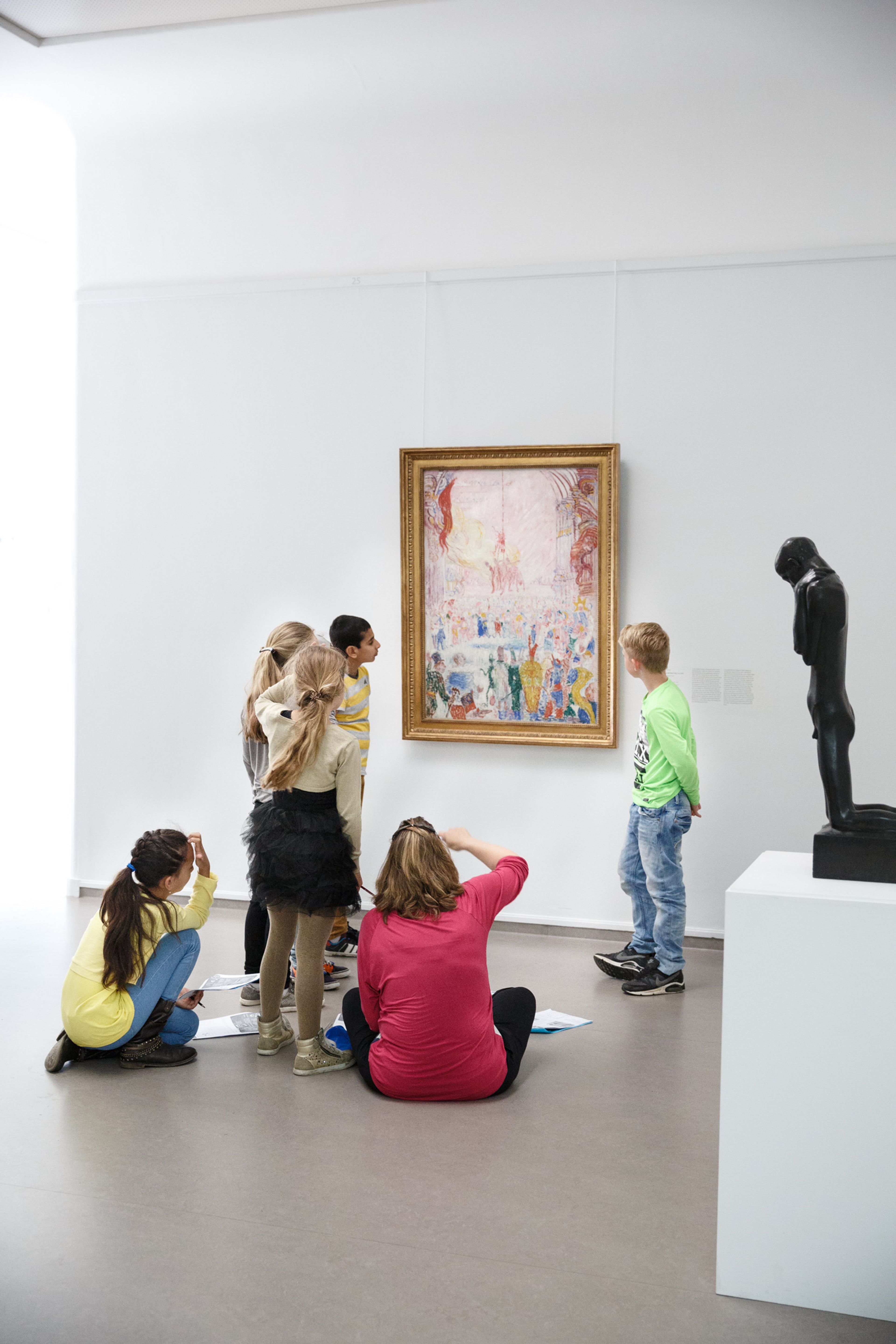 Groep kinderen bekijkt en bestudeert een schilderij in een museumruimte met witte muren en een grijze vloer. Links zitten drie kinderen op de grond, rechts staan drie kinderen, terwijl een zwarte beeldhouwwerk op een sokkel rechts in beeld staat.