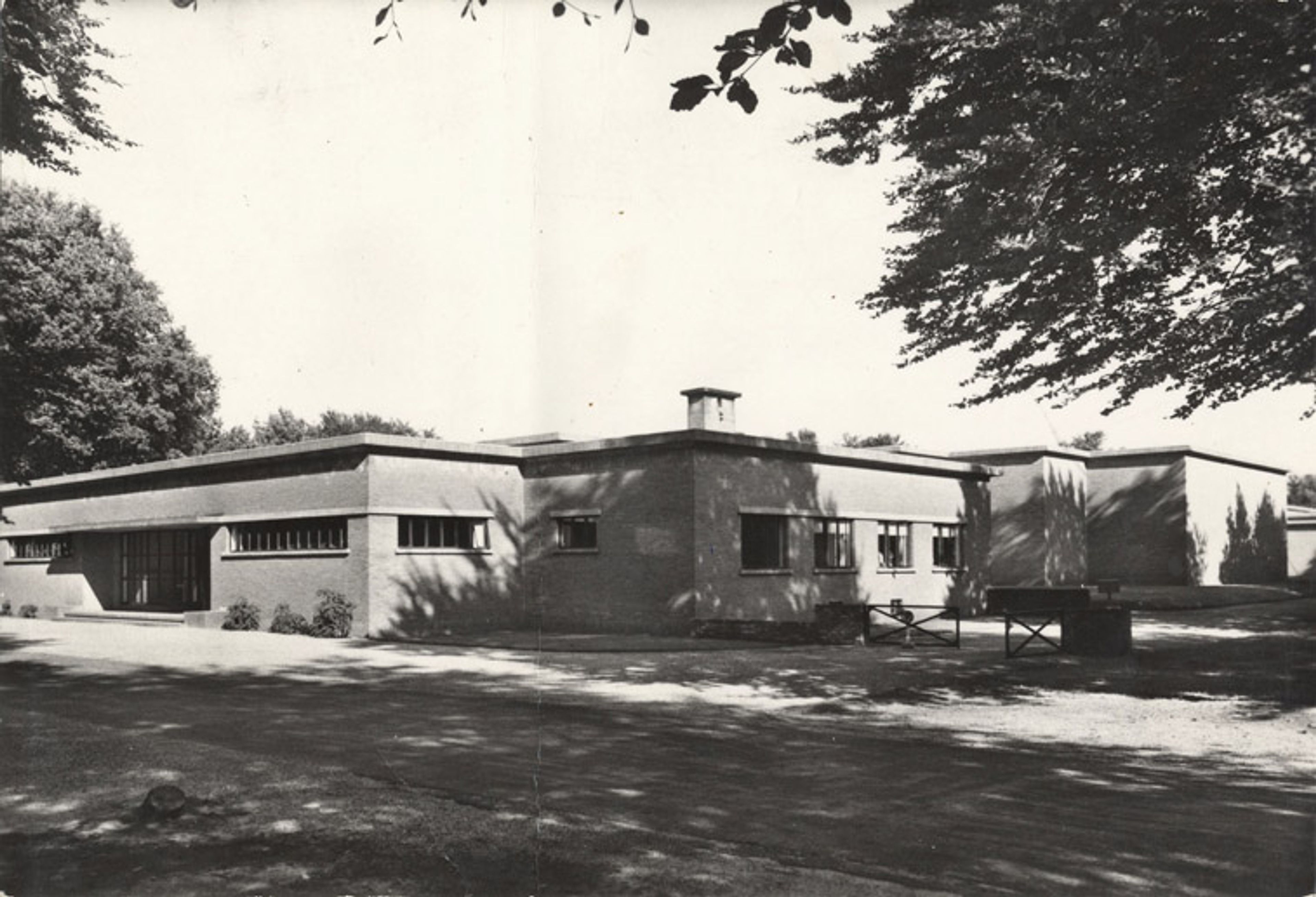 Het ‘Overgangsmuseum’ van Henry van de Velde Zwart-witfoto van het museum. Een laag, modern gebouw met strakke lijnen en platte daken, tussen bomen en omgeving met schaduw. Foto benadrukt de architectuurstijl en rustige, natuurlijke setting zonder mensen of auto’s.