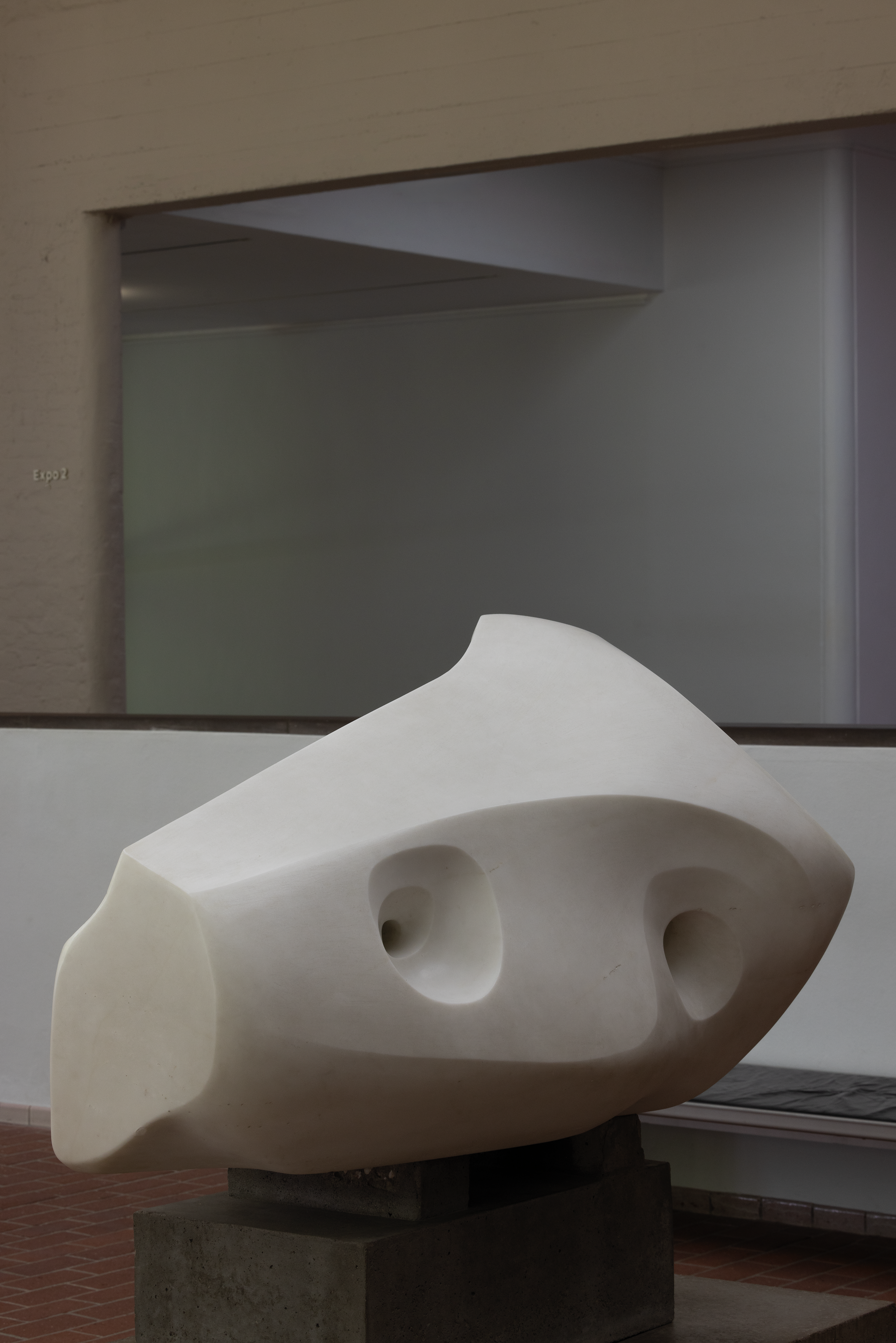 Foto van een abstracte, witte sculptuur met afgeronde vormen en twee ovale openingen, geplaatst op een donkere sokkel in een binnenruimte met bakstenen vloer.