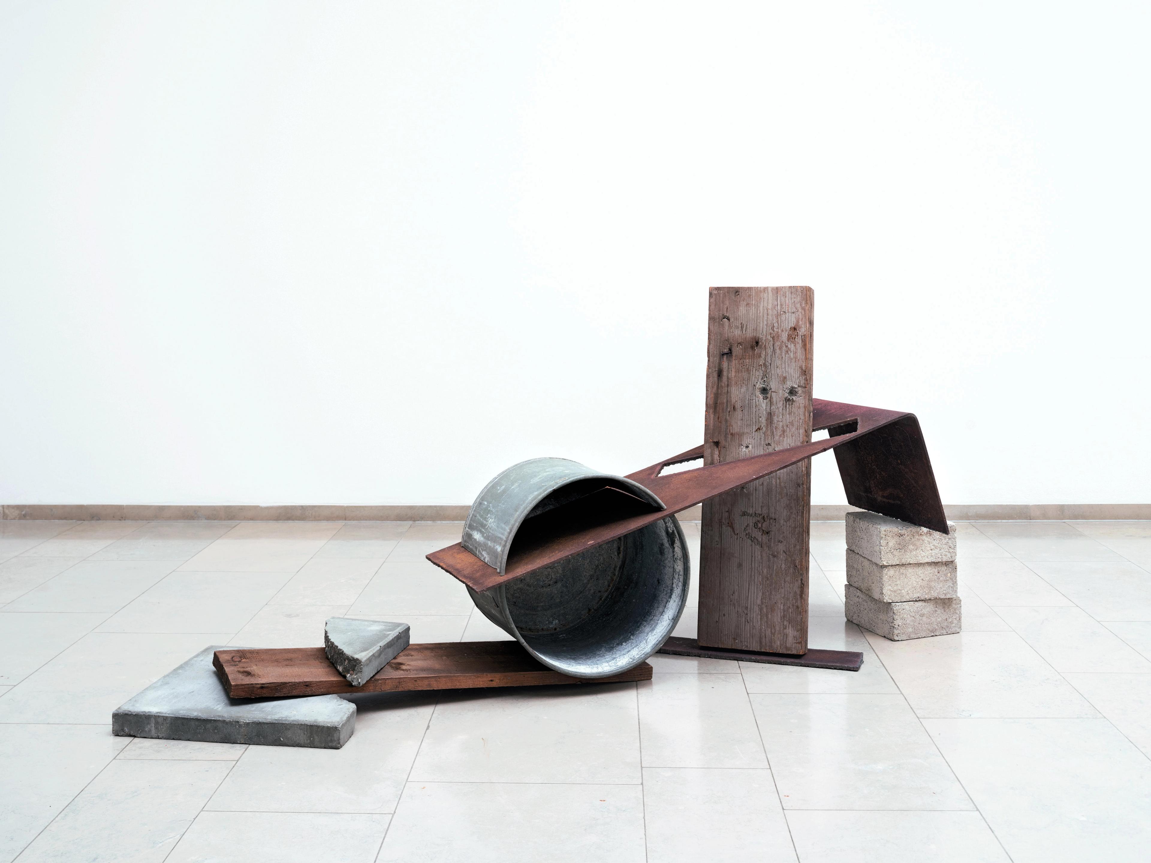 Ruud Kuijer, _Sculptuur I_, 1990, fotograaf Rüdiger Lubricht Foto van een abstracte sculptuur bestaande uit metalen platen, houten balken en blokken op een lichte tegelvloer tegen een witte muur.