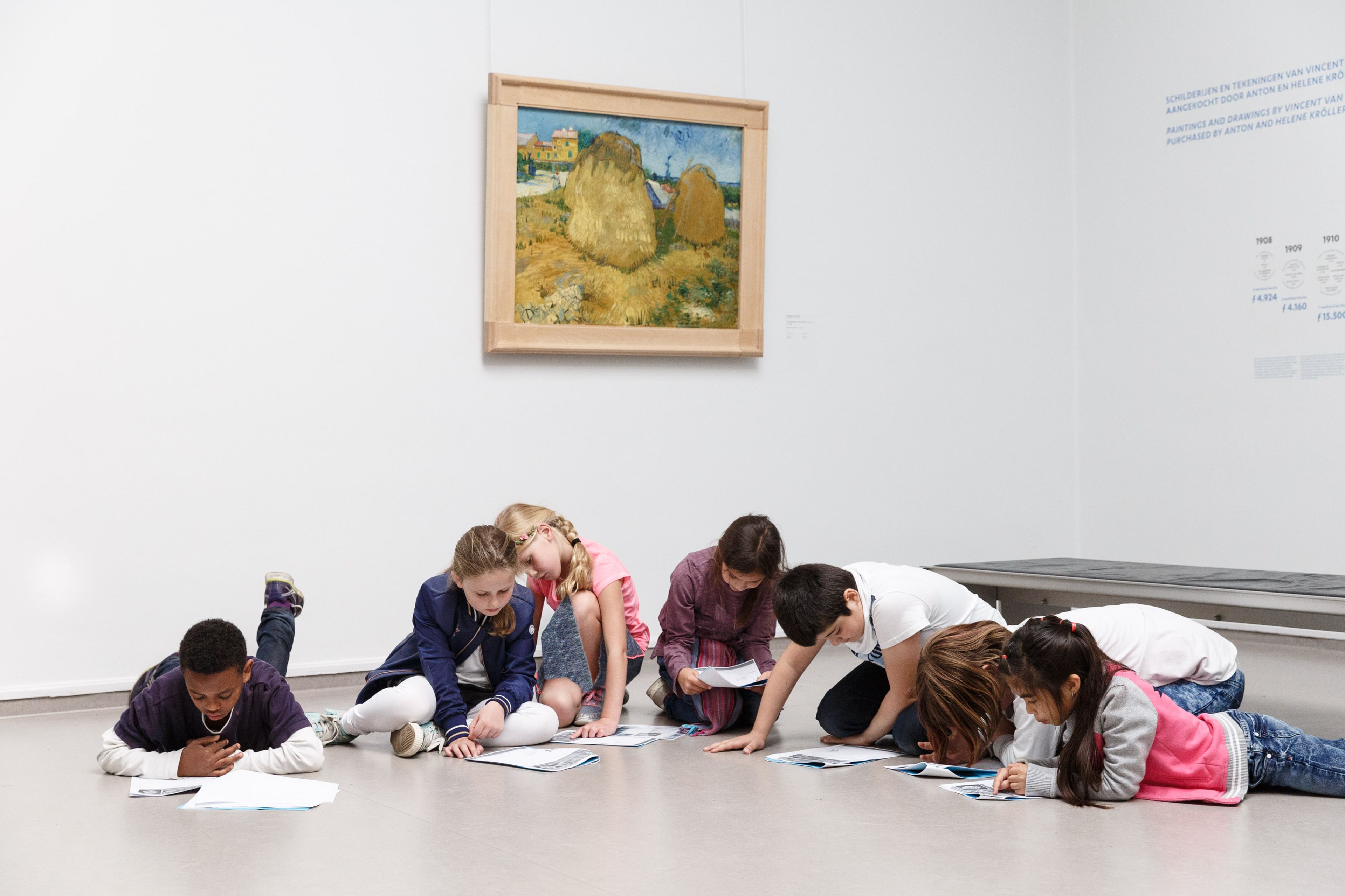 Groep kinderen die op vloer van museumzaal zitten en bezig zijn met tekenen of schrijven. Aan muur hangt schilderij met landschap en hooi, omgeving is minimalistisch met witte muren en informatiepanelen.