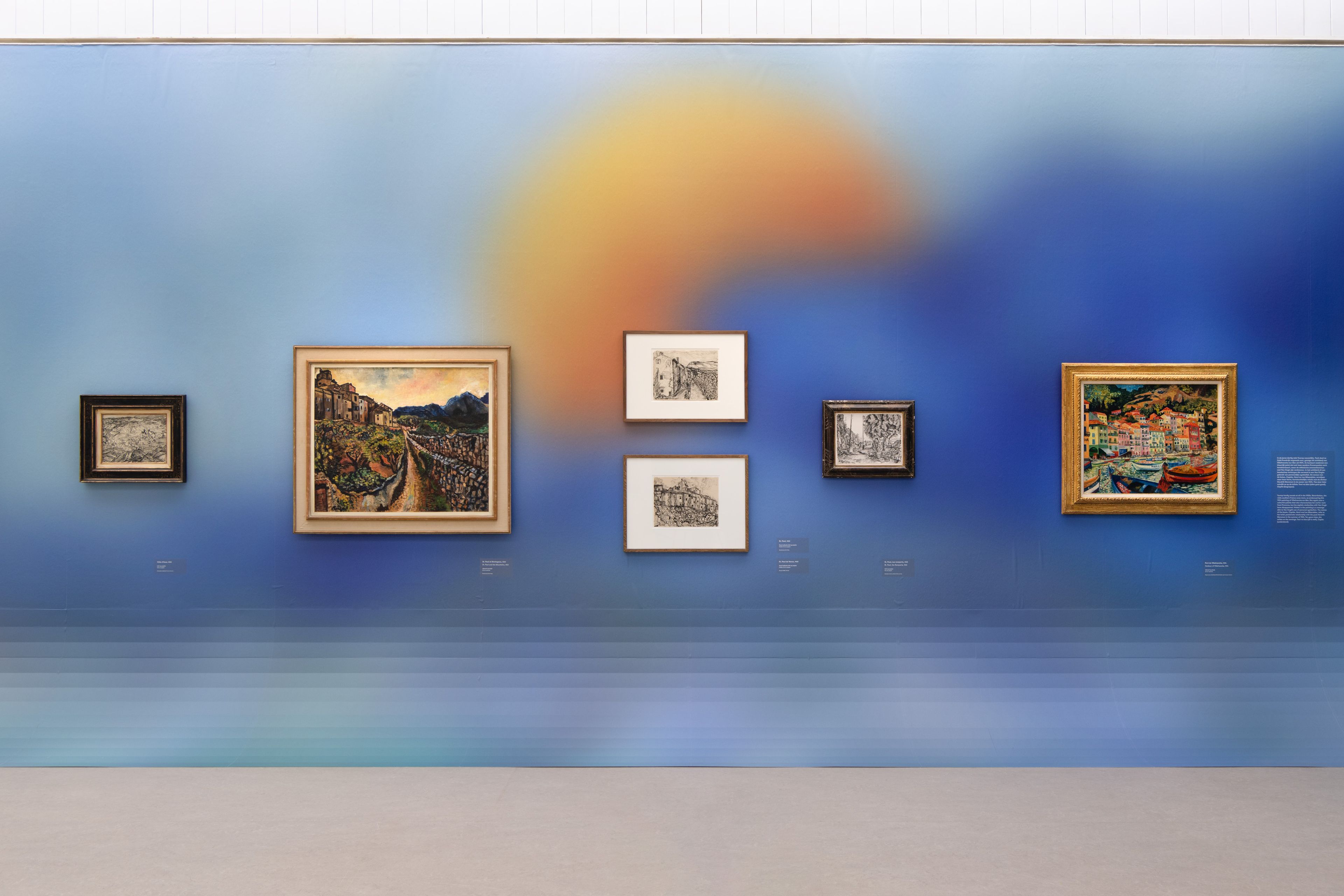 Zaaloverzicht _Charley Toorop. Liefde voor Van Gogh_ Foto van een wand met zes ingelijste schilderijen en tekeningen, geplaatst tegen een blauwe achtergrond met een grote, vervaagde oranje cirkel.
