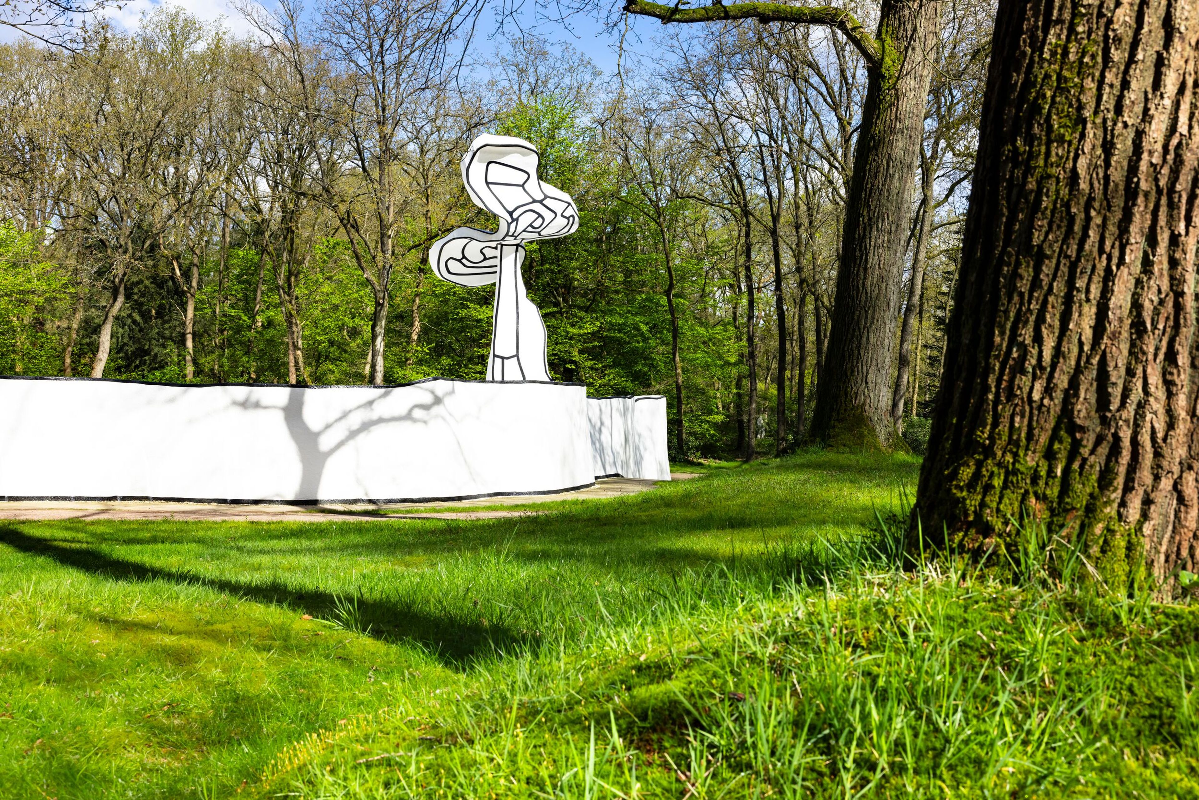 Foto van een buitenkunstwerk. Wit sculptuur met abstracte vormen staat op een verhoogd platform, omgeven door schaduwen van takken en heldere blauwe lucht.