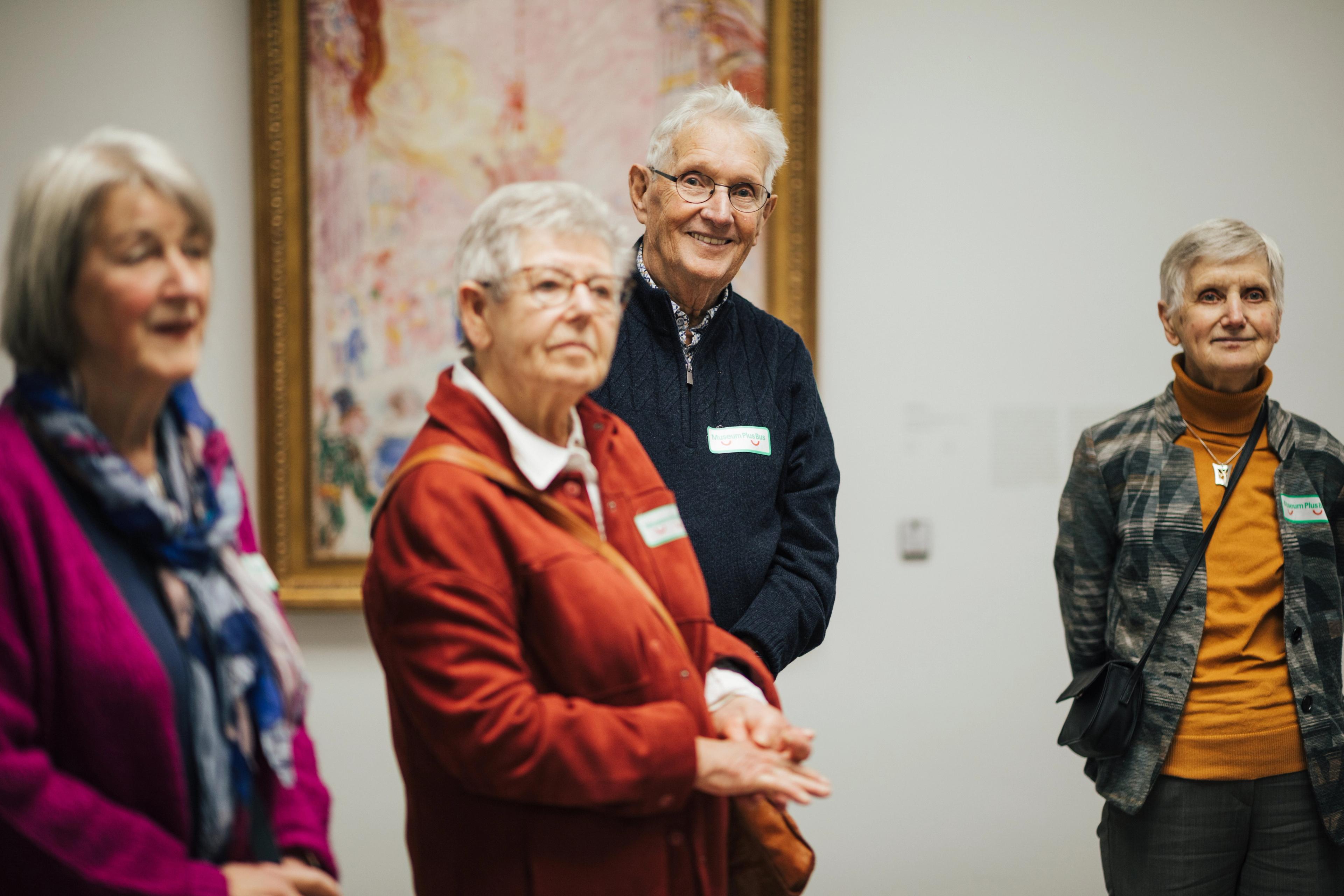 Foto van een groep volwassenen die in een museum staan, met een schilderij op de achtergrond.
