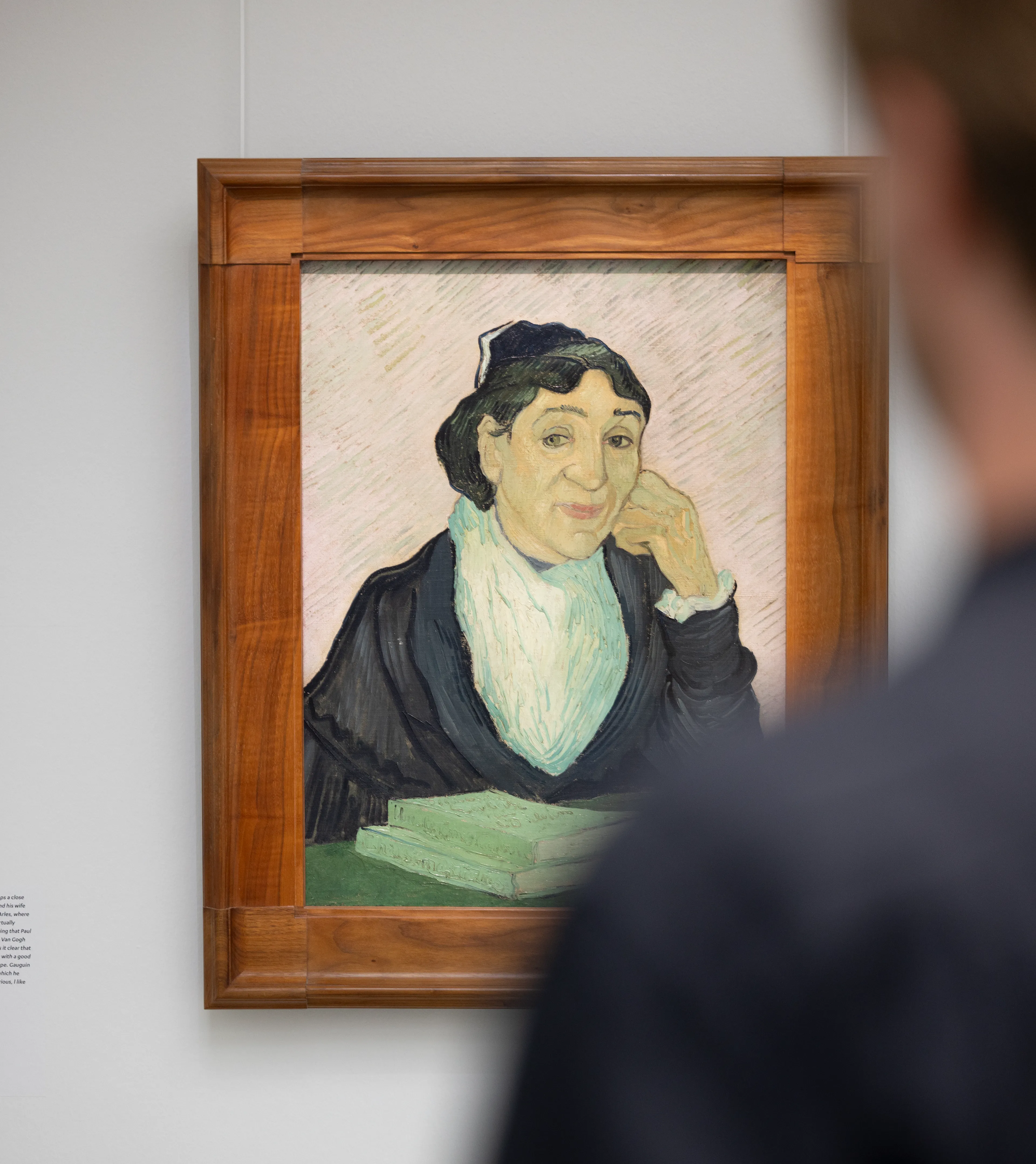 Photo of a person looking at a painting. The painting depicts a woman wearing dark clothing and a white scarf, framed by a wooden frame, with a book on the table in front of her.