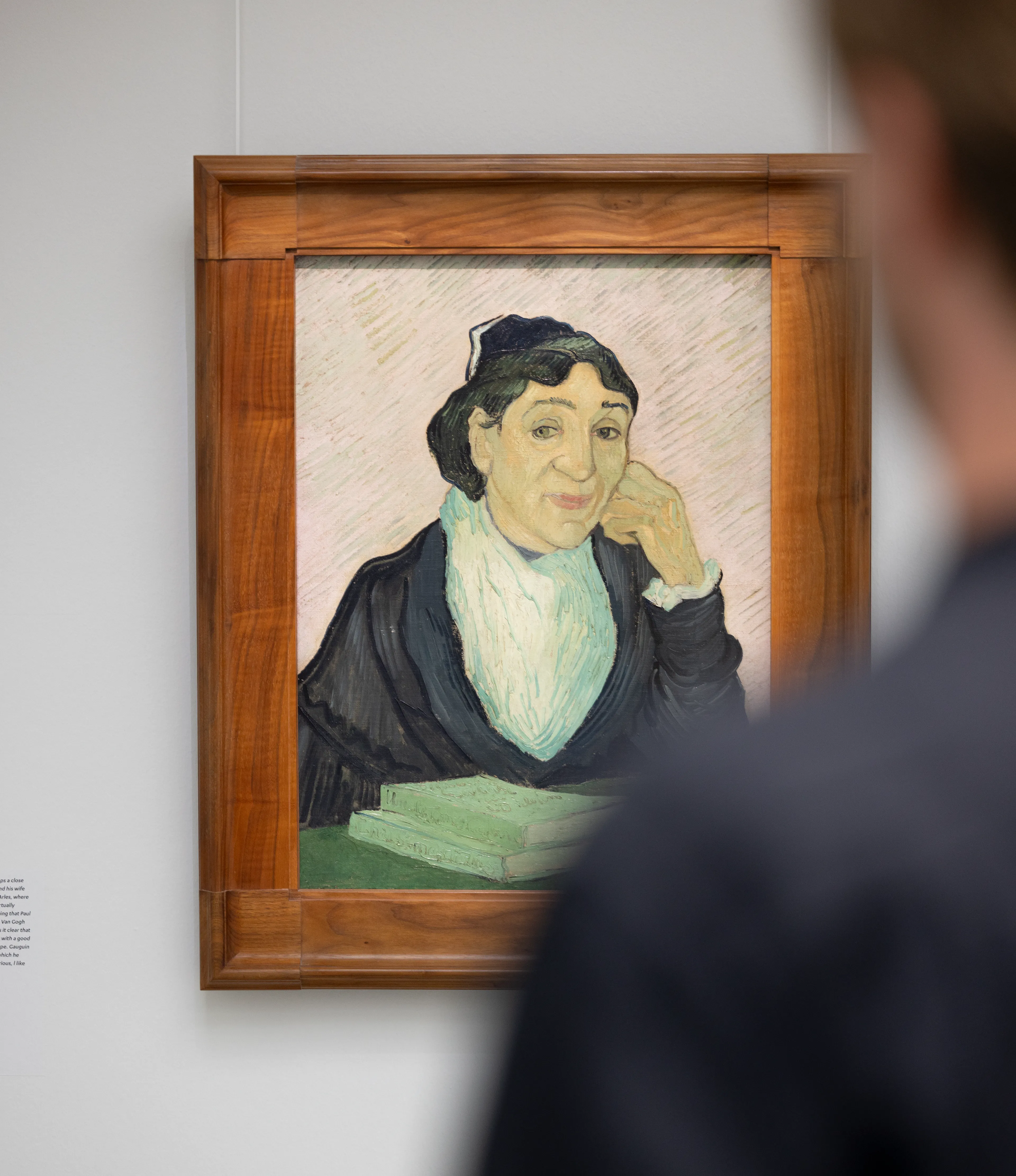 Foto van een persoon die naar een schilderij kijkt . Schilderij toont een vrouw met donkere kleding en een witte sjaal, omlijst door een houten lijst, met een boek op tafel voor haar.