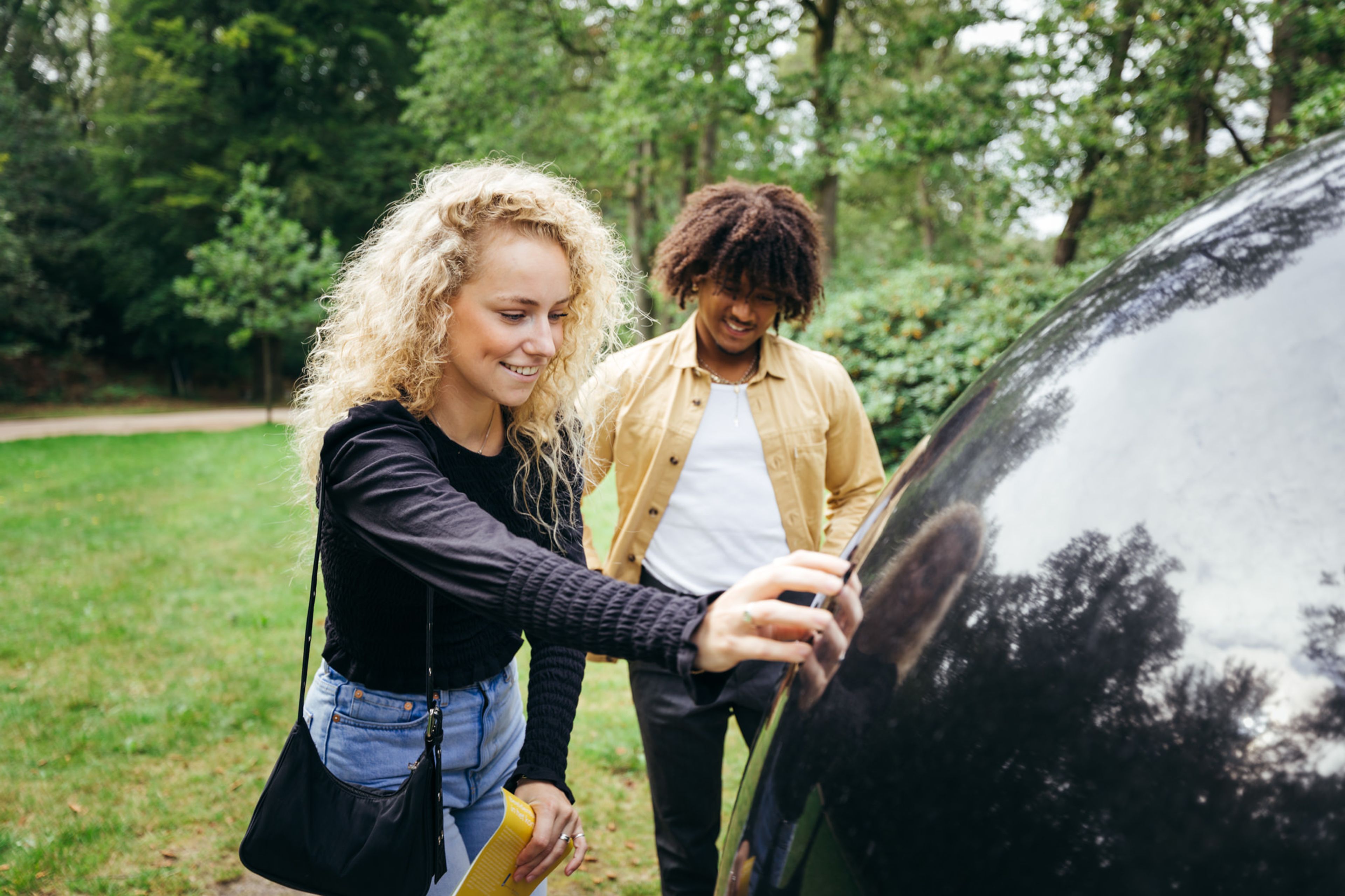 Twee jonge mensen staan buiten bij een groot, glanzend zwart object in een groene parkachtige omgeving. Een persoon raakt het object aan terwijl ze beiden aandachtig kijken, wat een moment van nieuwsgierigheid of ontdekking suggereert.