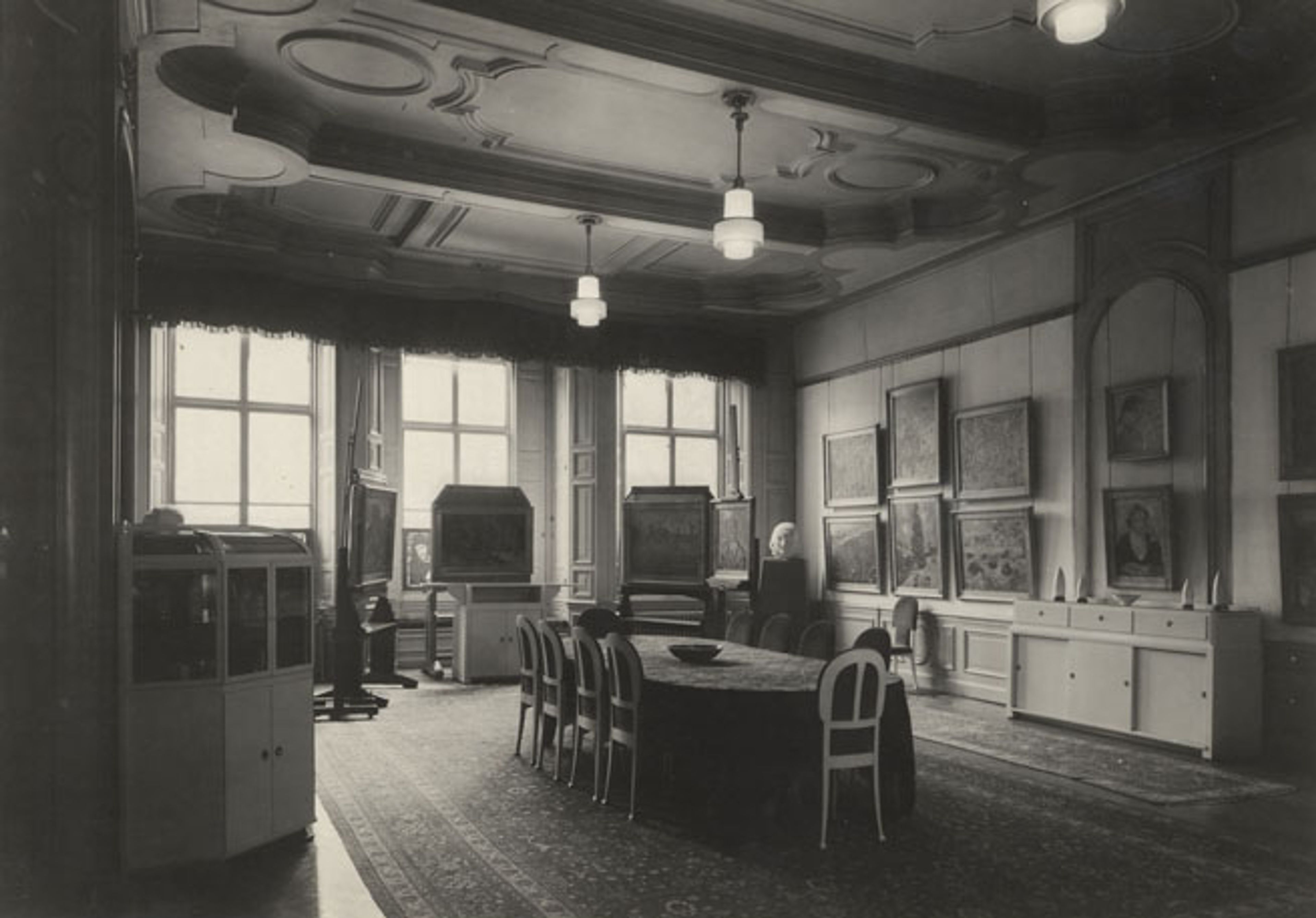 ‘Museum Kröller’ aan het Lange Voorhout Zwart-witfoto van een klassieke vergaderruimte met een lange tafel en stoelen. Aan de muren hangen schilderijen en grote ramen laten natuurlijk licht binnen. Kamer heeft bewerkt plafond, lampen en een tapijt op houten vloer, wat voor een formele en historische sfeer zorgt.