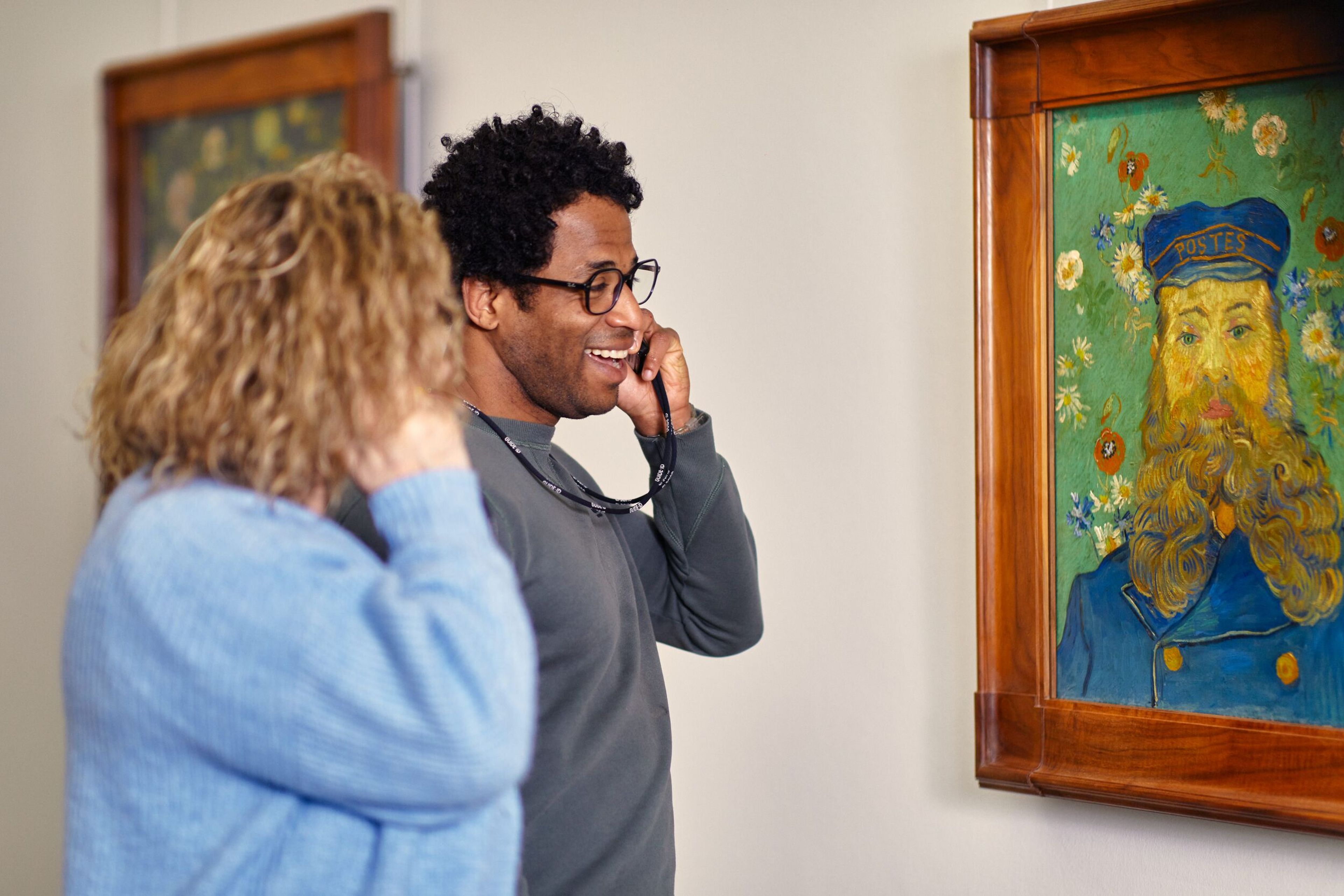 Twee personen bekijken een schilderij in een museum, waarbij ze beide een audiotour gebruiken. Schilderij toont een kleurrijk portret van een persoon met een blauwe pet en een baard, omlijst met een houten lijst.