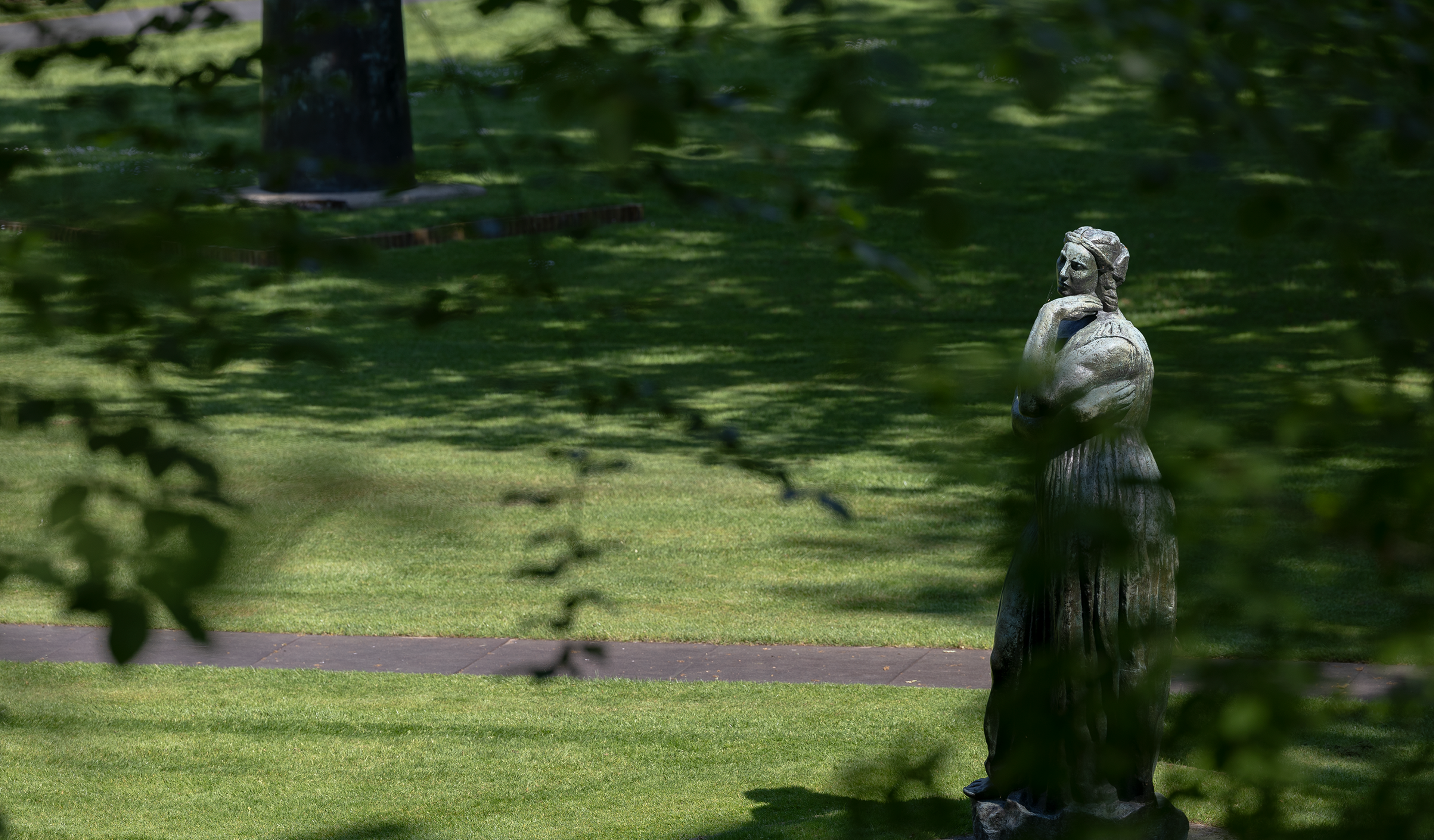 Émile-Antoine Bourdelle, _La grande Pénélope_, 1912 Foto van een beeld op groen gras en schaduwen van bomen op de achtergrond. Beeld toont een persoon met armen over elkaar,