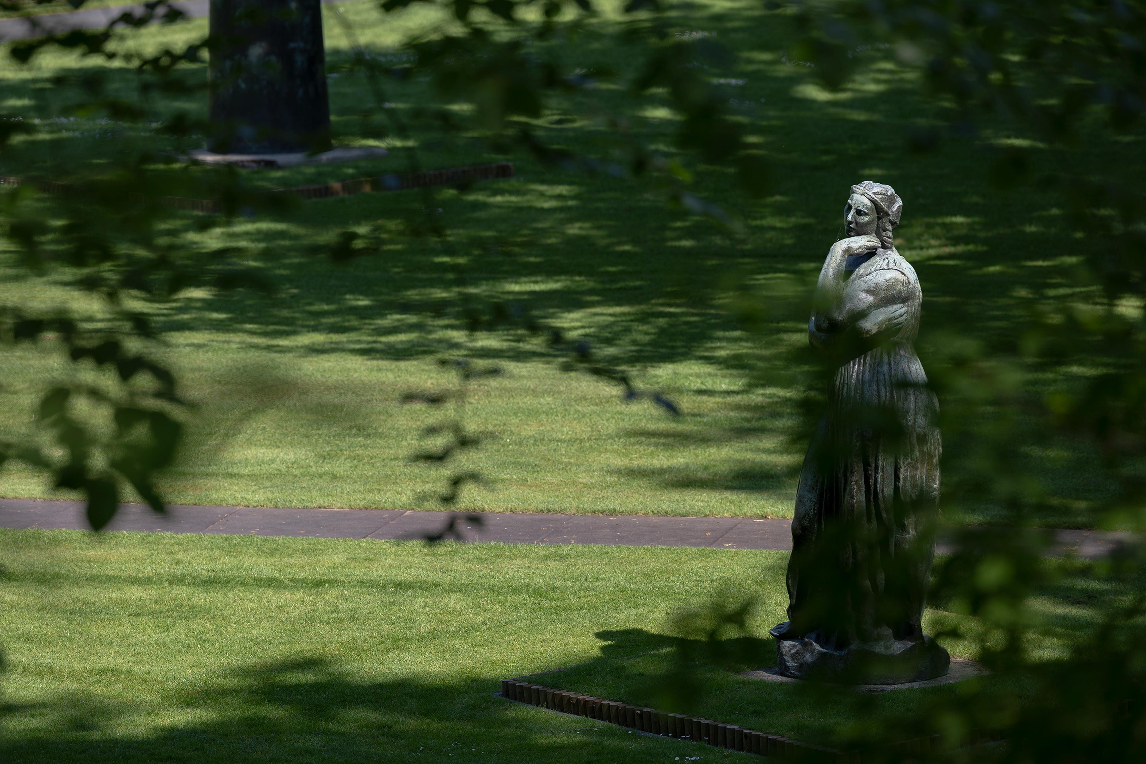 Émile-Antoine Bourdelle, _La grande Pénélope_, 1912 Foto van een beeld op groen gras en schaduwen van bomen op de achtergrond. Beeld toont een persoon met armen over elkaar,