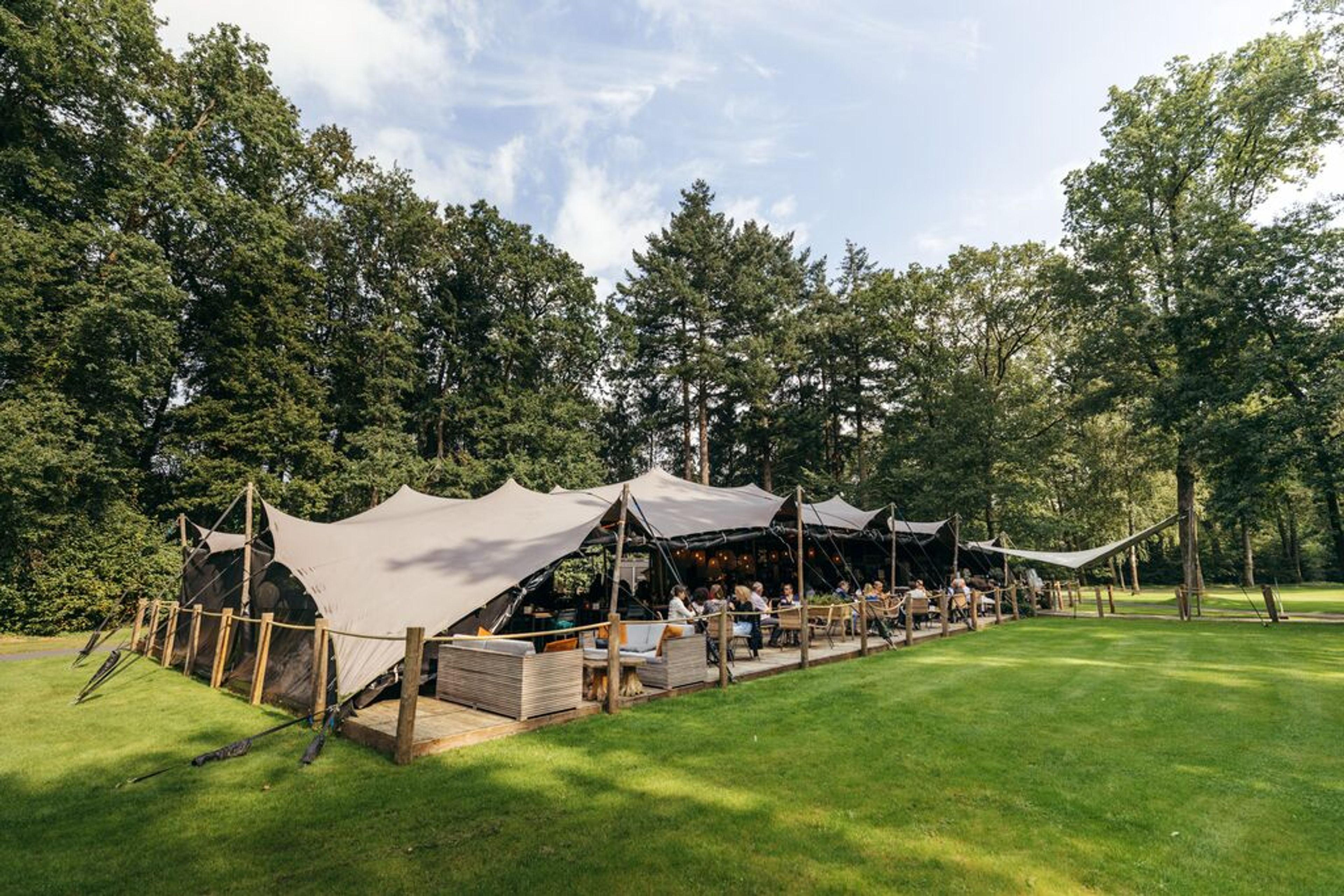 Foto van een restaurant buiten tussen het groen. Mensen op het terras en gedeeltelijk in de schaduw onder een doek