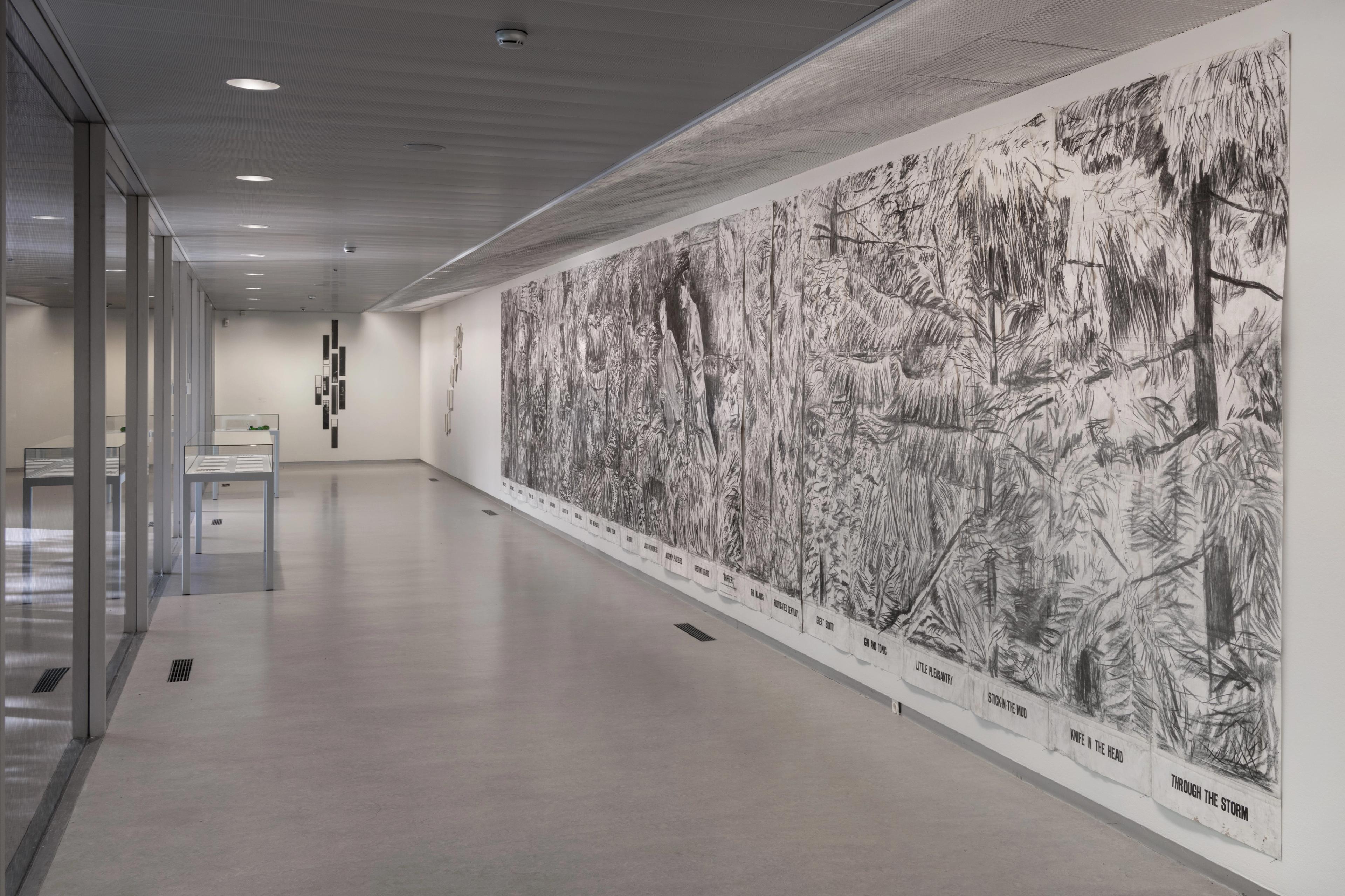 Grote zwart-witte wandtekening in een museumruimte, met twee figuren in een dicht begroeid landschap; de tekening loopt over meerdere panelen langs een lange witte muur in een tentoonstellingszaal.
