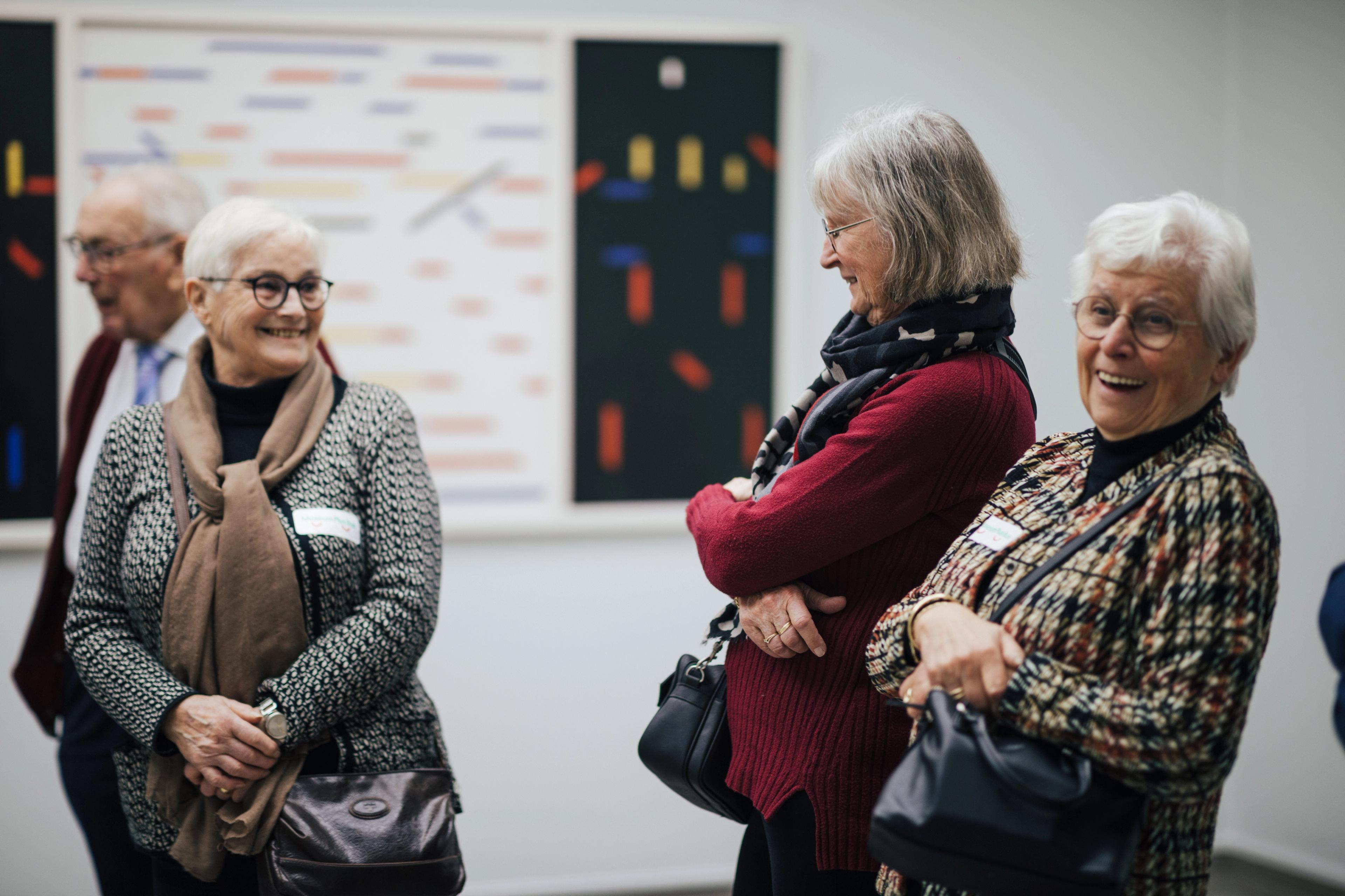Foto van vier oudere personen die een museum bezoeken, met abstracte schilderijen op de achtergrond.