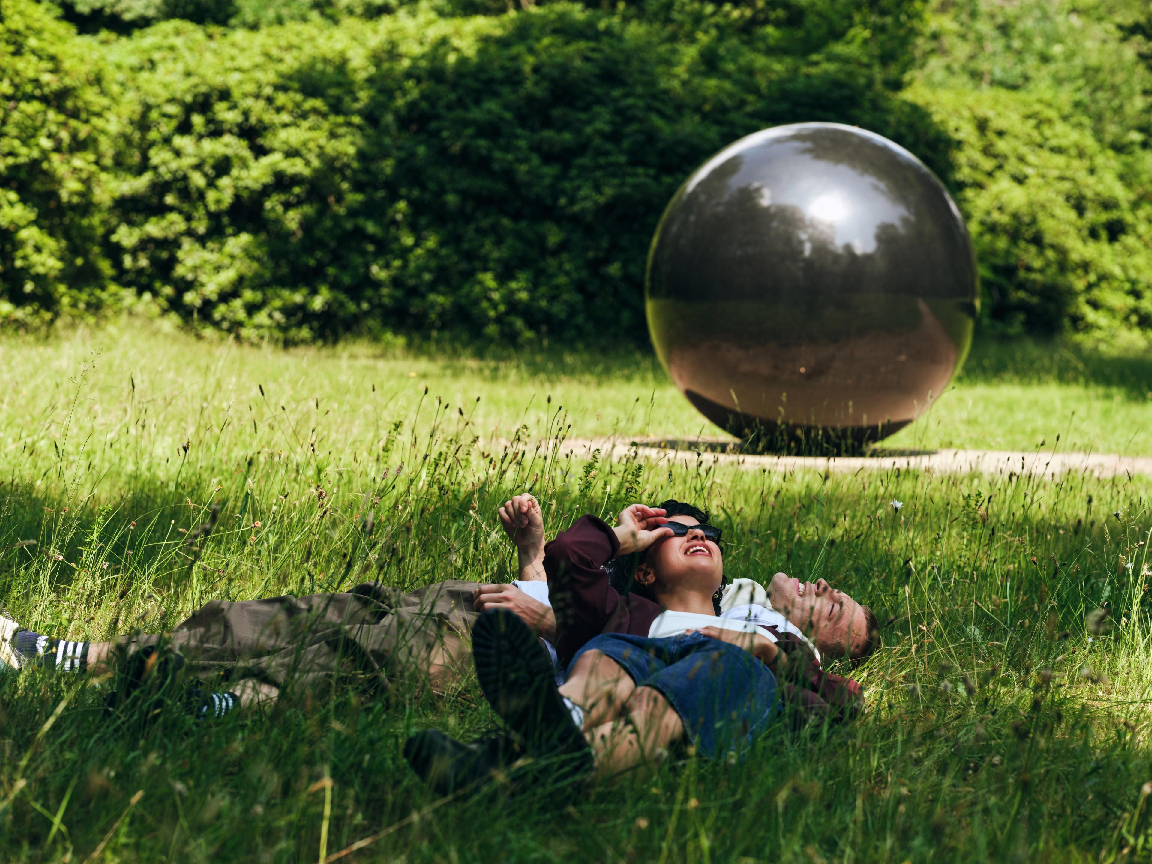 Foto van twee personen die ontspannen liggen in grasveld met groene bomen op achtergrond en kunstwerk, een grote, glanzende zwarte bol 
