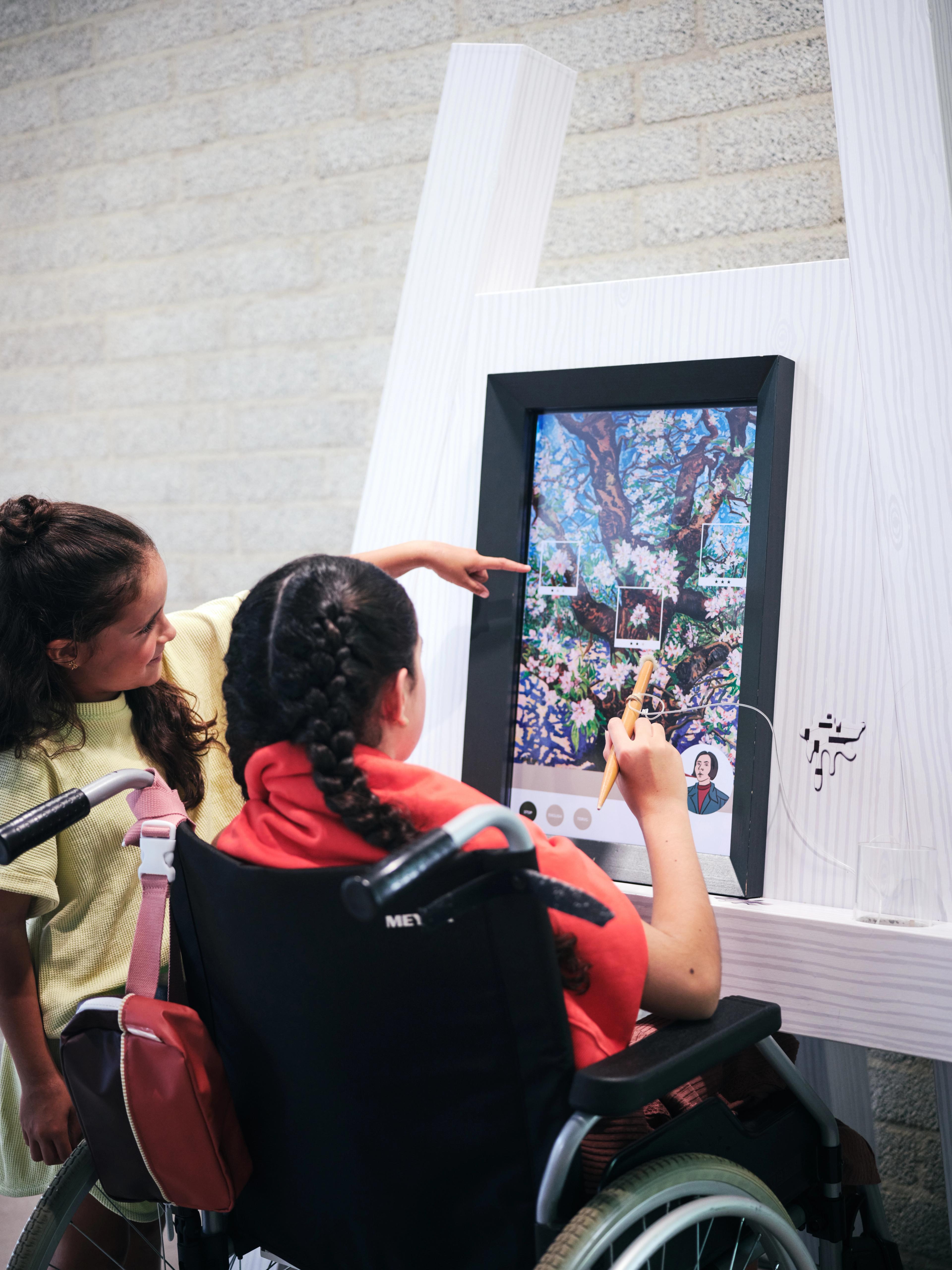 Foto van twee kinderen die een interactief touchscreen gebruiken; een kind in een rolstoel bedient het scherm met een pen terwijl de ander toekijkt.
