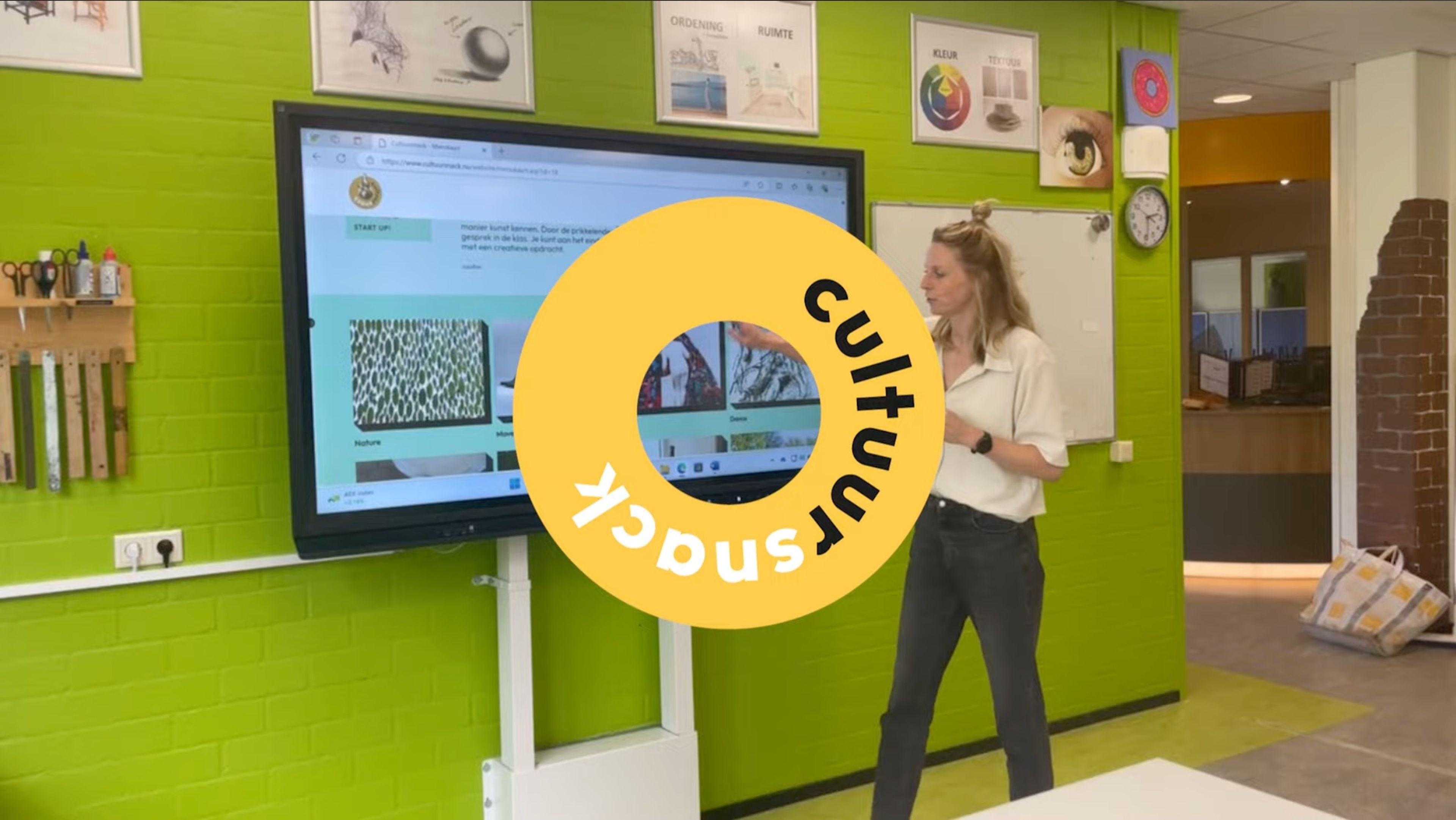 Foto van een klaslokaal met een groot interactief scherm aan een groene muur, waar een vrouw voor staat en iets presenteert. Op het scherm is een website zichtbaar en in het midden een geel-oranje cirkel met tekst "cultuursnack" in zwarte letters.