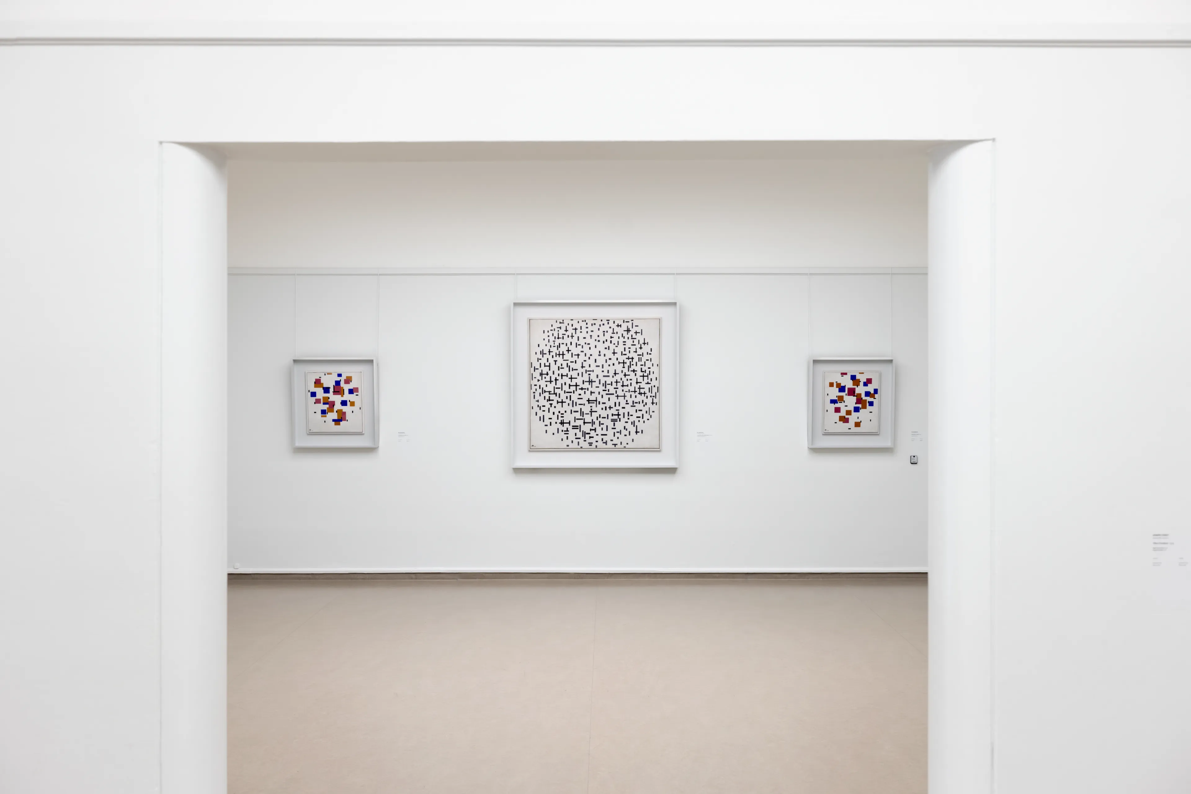 Foto van een zaal in het Kröller-Müller Museum, witte wanden met een doorkijkje. Aan de muur hangen drie schilderijen van Piet Mondriaan. In het midden een groot schilderij in zwart wit, en twee schilderijen met kleur aan beide zijden. 