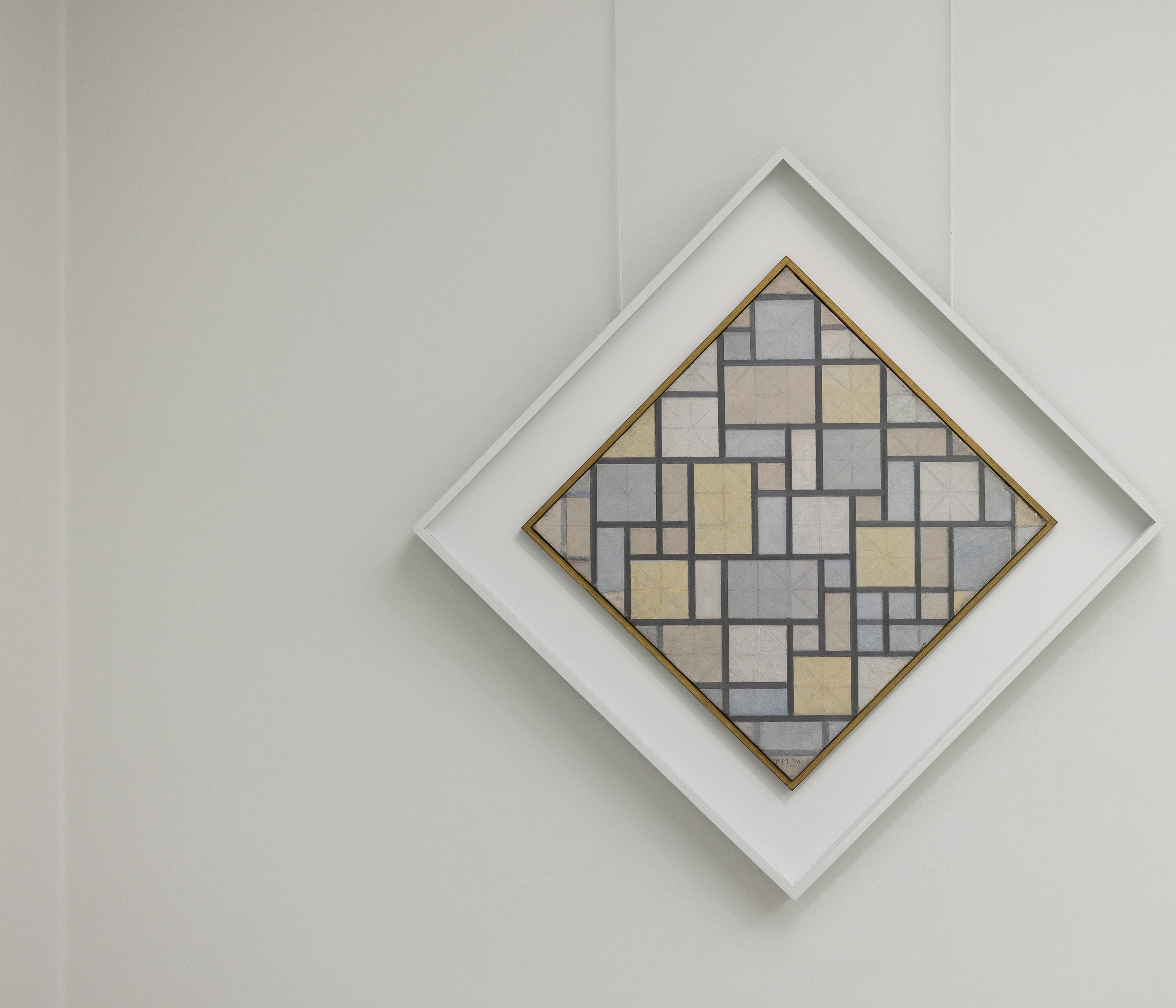 Foto van een schilderij van Piet Mondriaan bestaande uit een vierkant met een patroon van rechthoeken en vierkanten in grijs- en beige tinten, omlijst met een dunne gouden rand. Kunstwerk hangt diagonaal geplaatst in een witte, vierkante lijst tegen een witte muur.