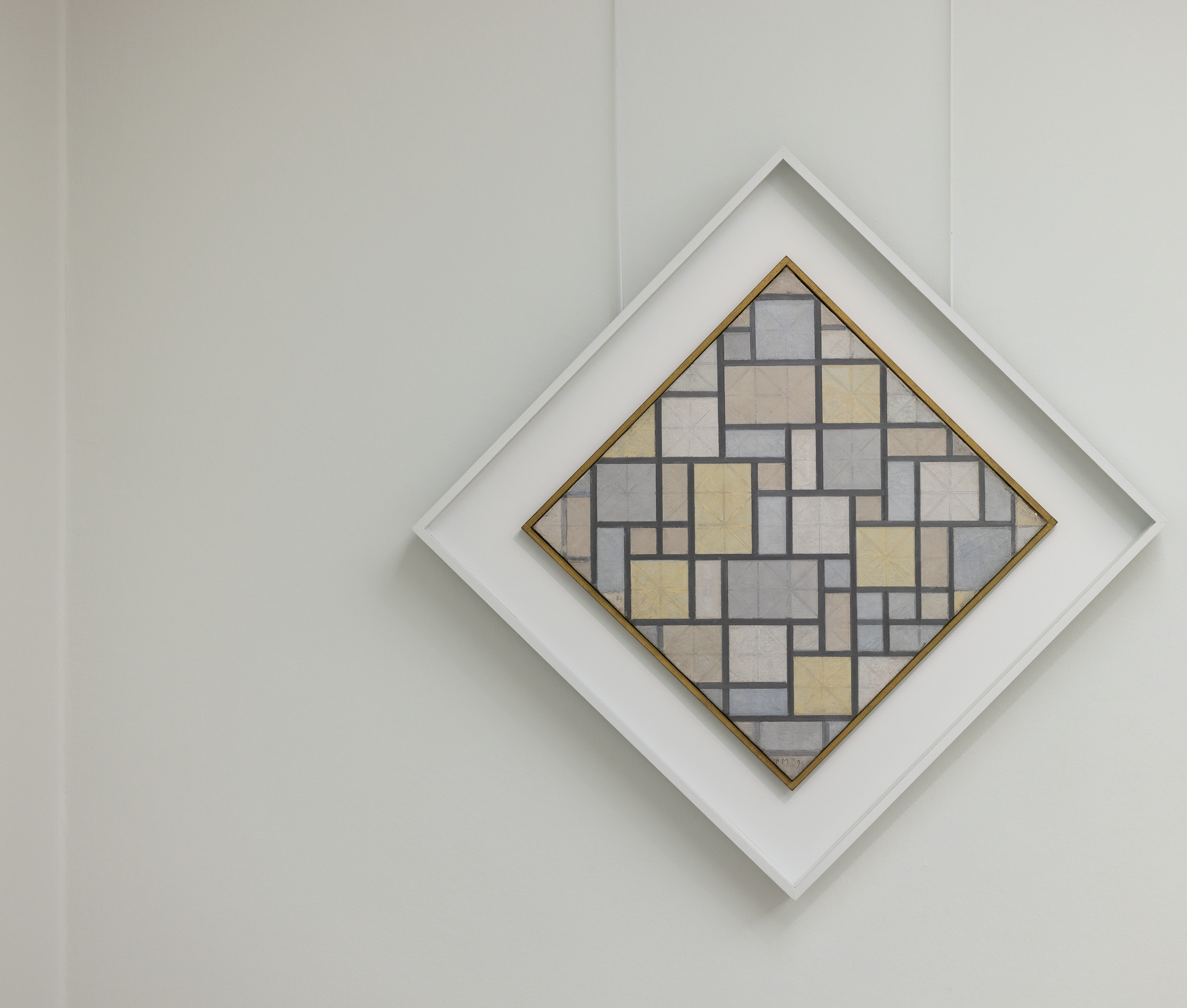 Photograph of a painting by Piet Mondrian consisting of a square with a pattern of rectangles and squares in shades of grey and beige, framed by a thin gold border. The artwork is hung diagonally in a white, square frame against a white wall.
