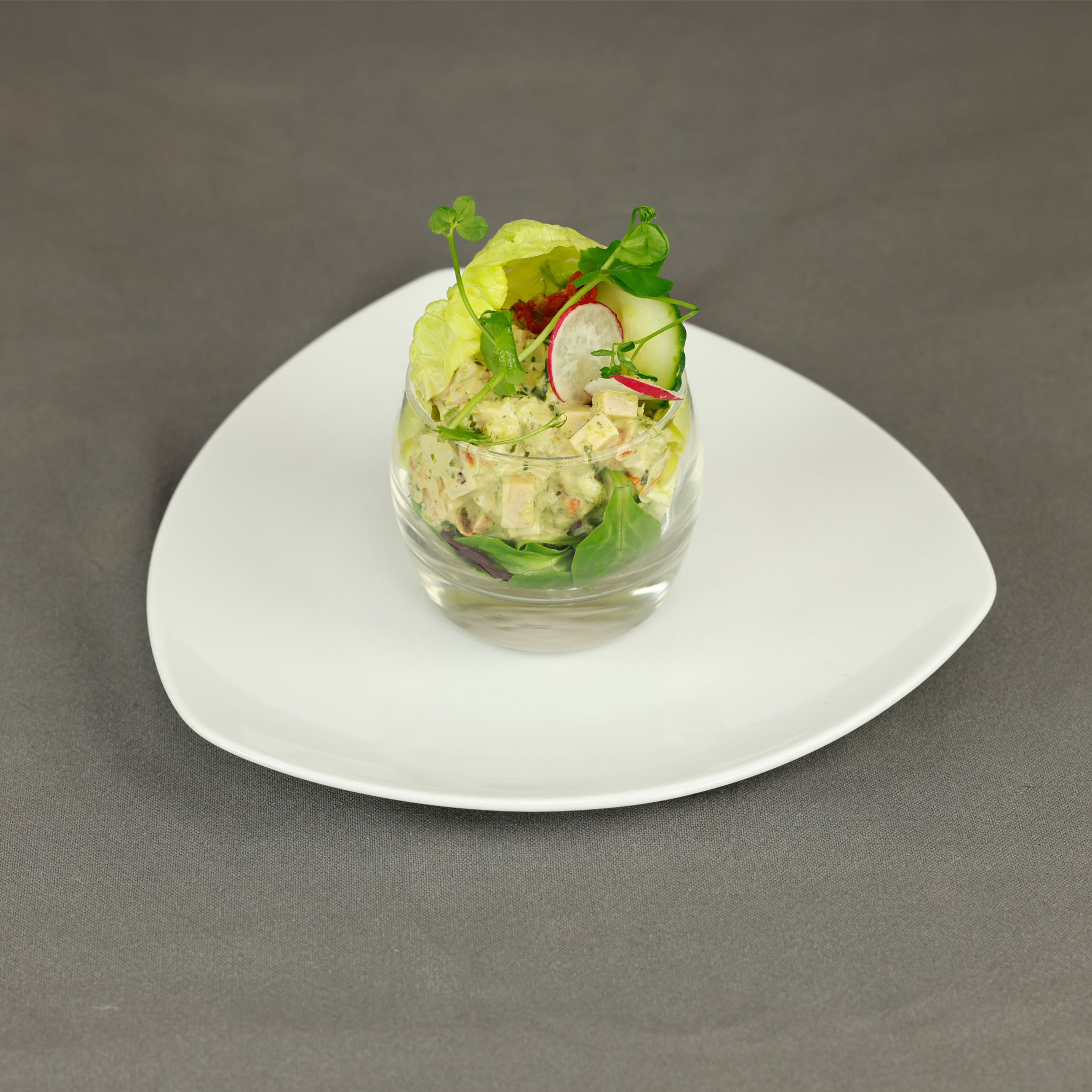 Photograph of a gourmet appetizer presented in a small glass on a white, triangular plate. The dish features a creamy salad with pieces of vegetables, garnished with lettuce leaves, radishes, and fresh herbs, set against a gray background.