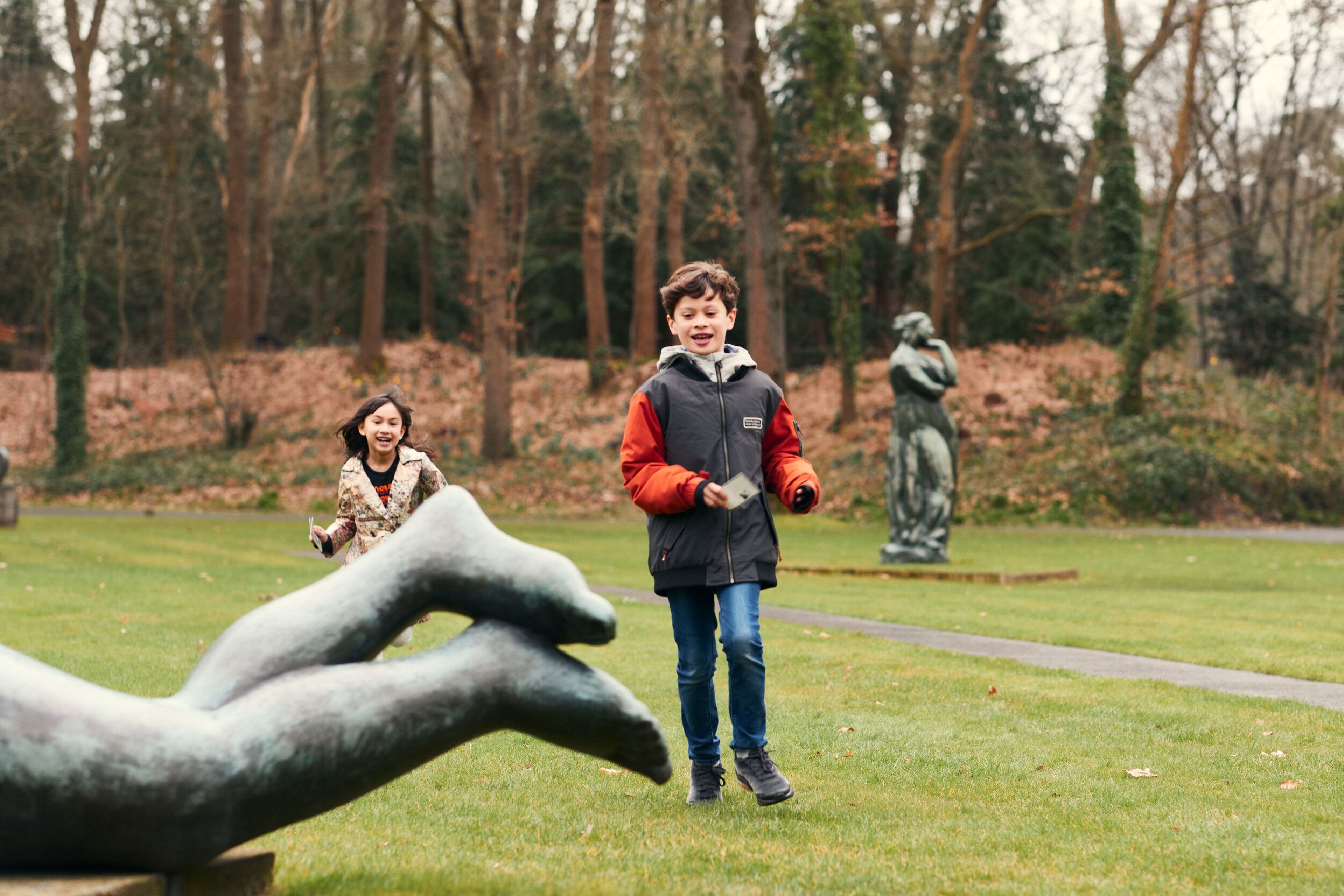 Foto van twee kinderen die spelen in de beeldentuin. Op de achtergrond een staand bronzen beeld en op de voorgrond zijn de benen van een liggende figuur te zien in brons. Het is een herfstachtige omgeving met kale bomen en een groen grasveld. 