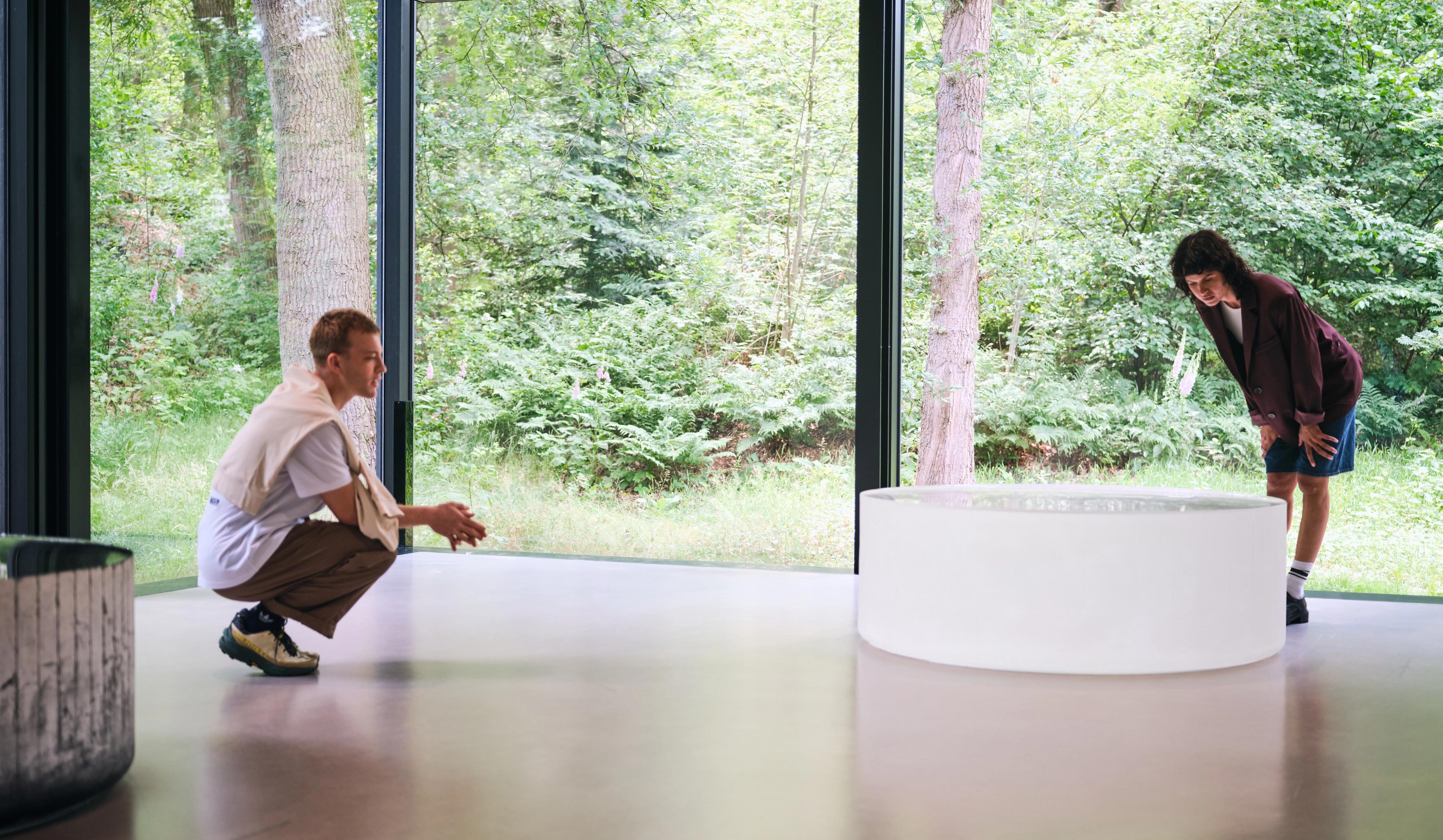 Twee mensen kijken naar een wit rond kunstwerk in gang van het museum. Door de grote glazen wanden zijn groene bomen en struiken te zien.