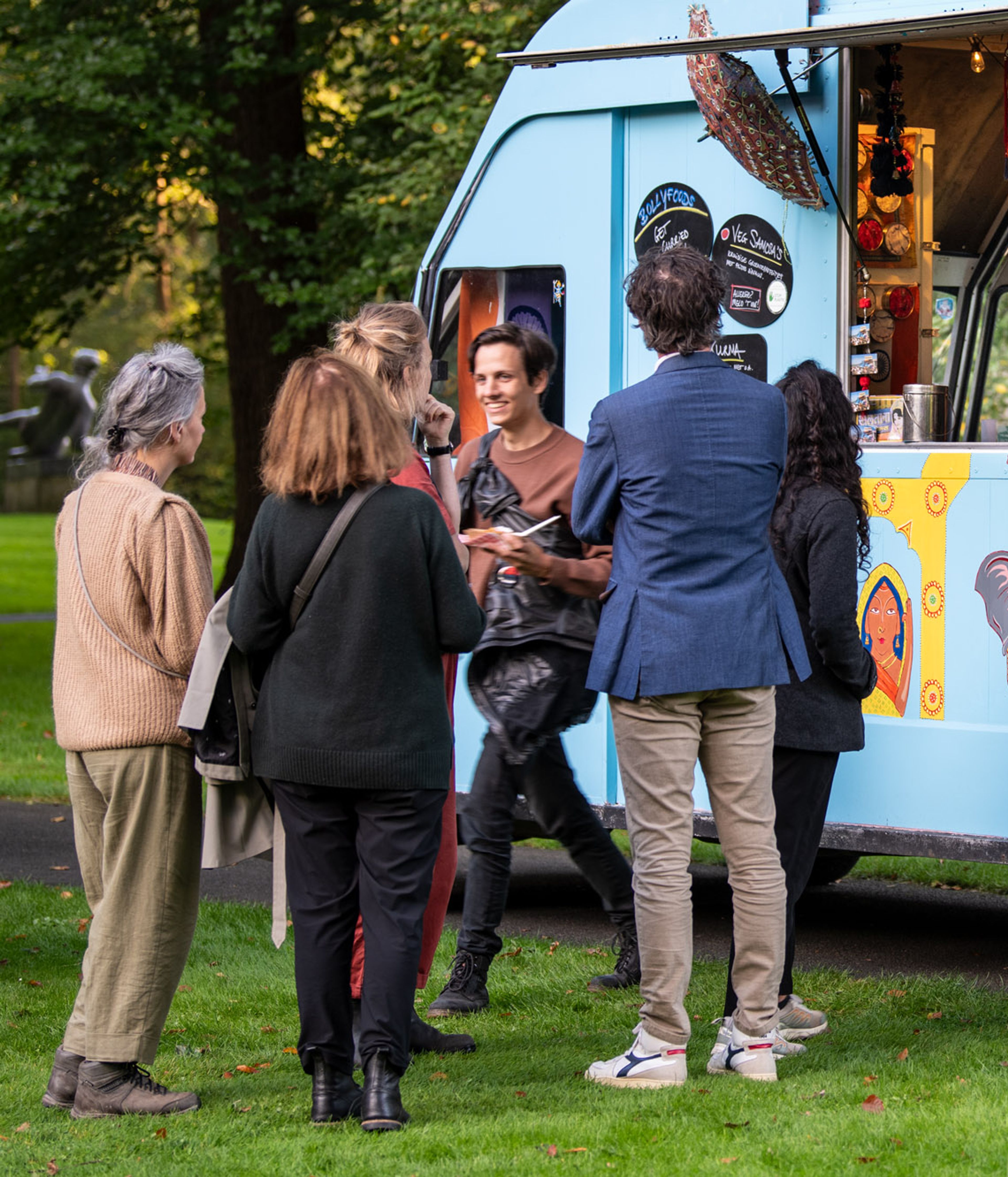 Foto van een gezelschap dat in de beeldentuin onstpannen met elkaar praat voor een foodtruck in frisse kleuren blauw