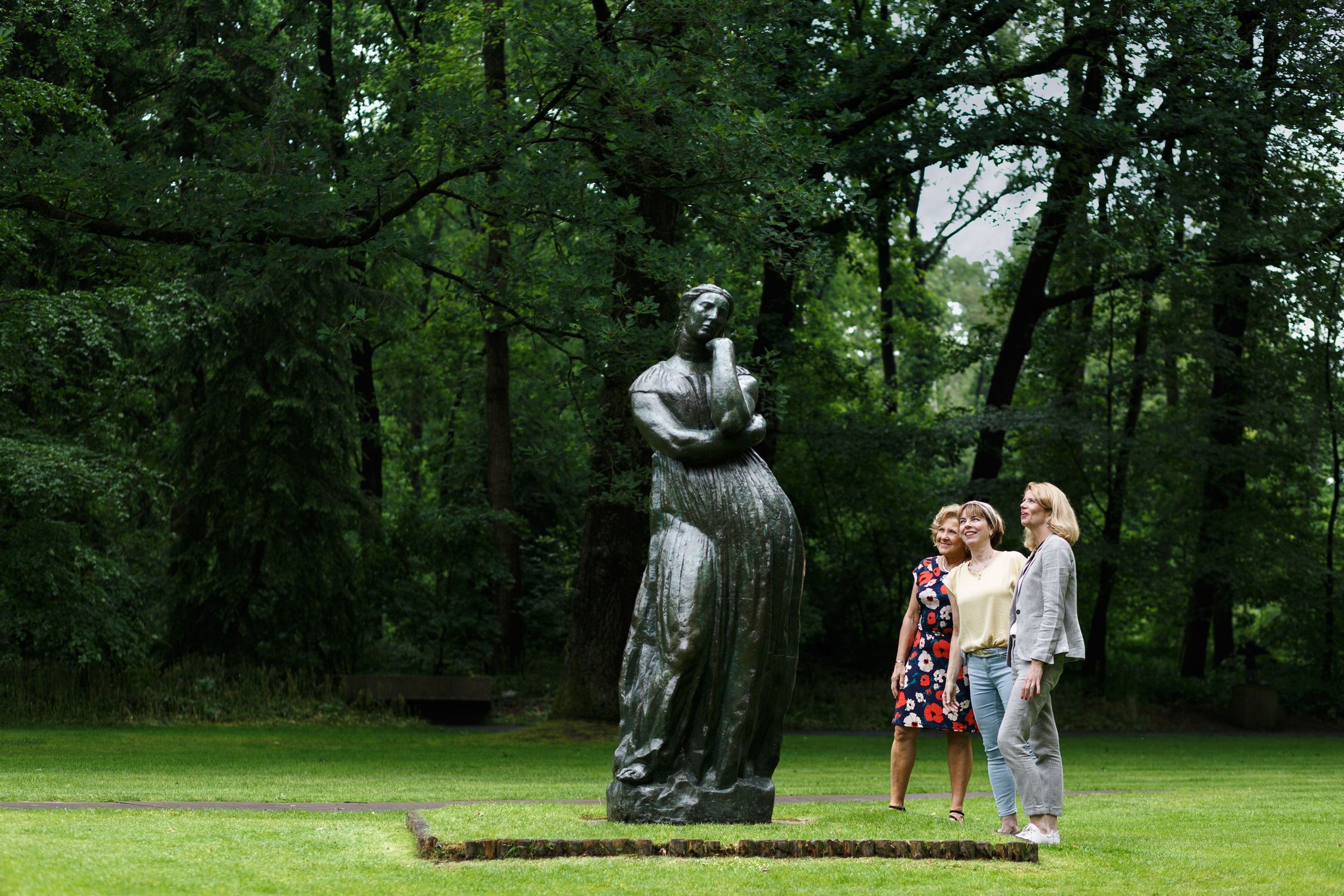 Foto van drie vrouwen die een groot, donker bronzen standbeeld van een vrouw in een park met groene bomen en gras bekijken. Het beeld toont een vrouw met gekruiste armen en lange jurk, terwijl vrouwen ernaast ontspannen en geïnteresseerd lijken.