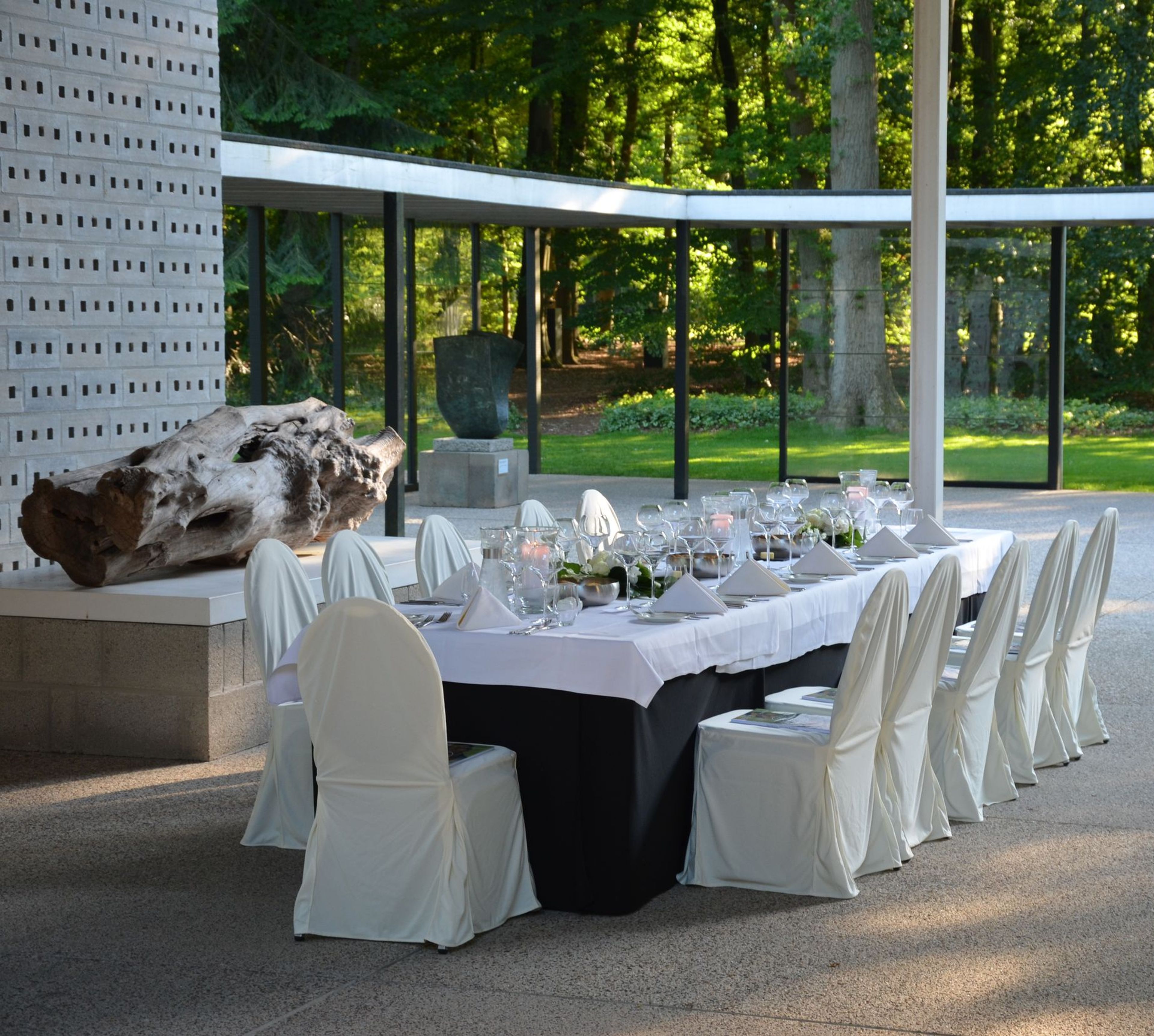 Gedekte lange eettafel met wit linnen en stoelhoezen in het Rietveldpaviljoen van het Kröller-Müller Museum, met servies en glaswerk klaar voor een diner; rondom glazen wanden met uitzicht op een groen bos en enkele sculpturen.