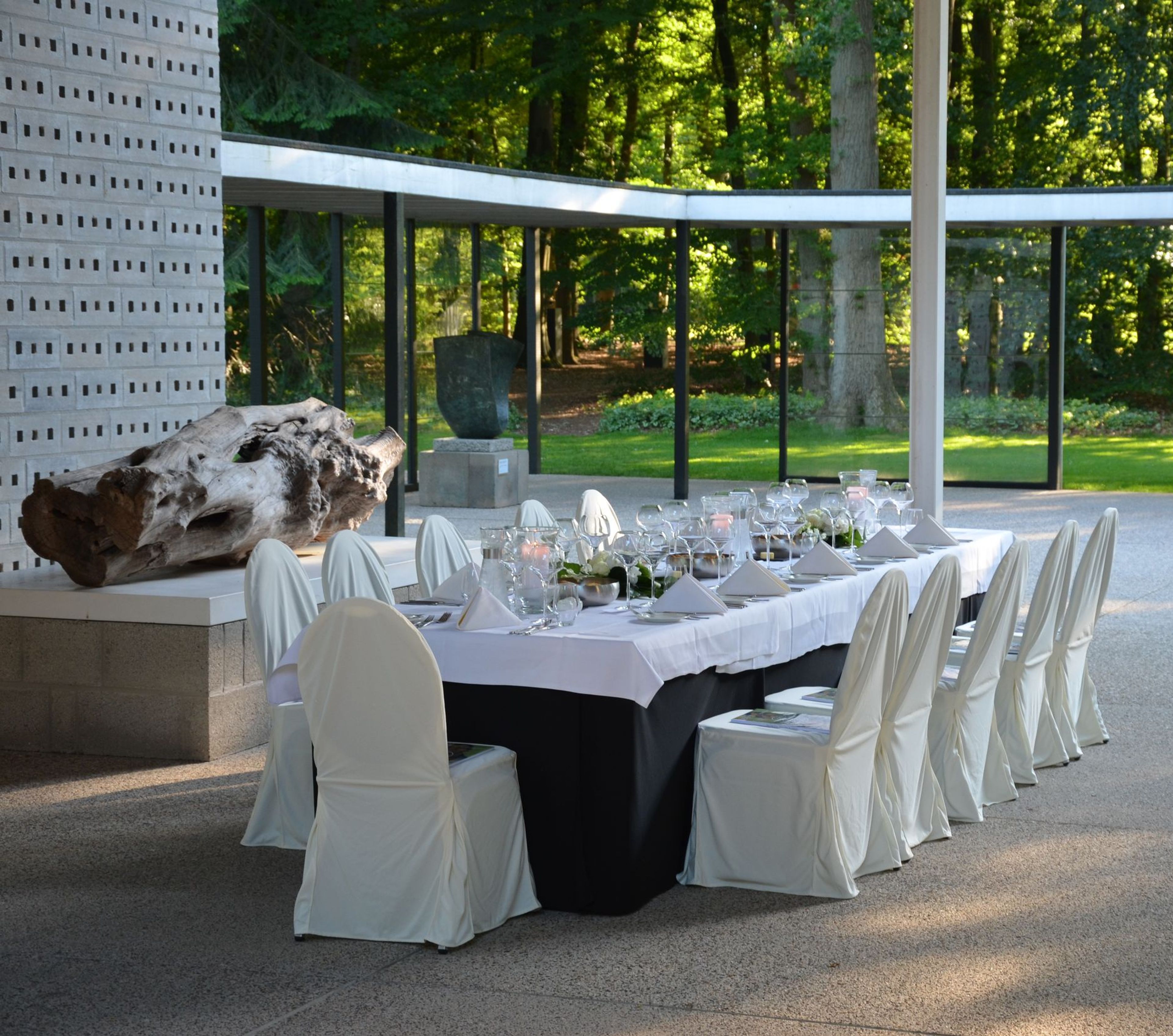 Long dining table set with white linen and covered chairs inside the Rietveld Pavilion at the Kröller-Müller Museum, with tableware and glassware arranged for a dinner; glass walls offer views of the surrounding forest and sculptures.