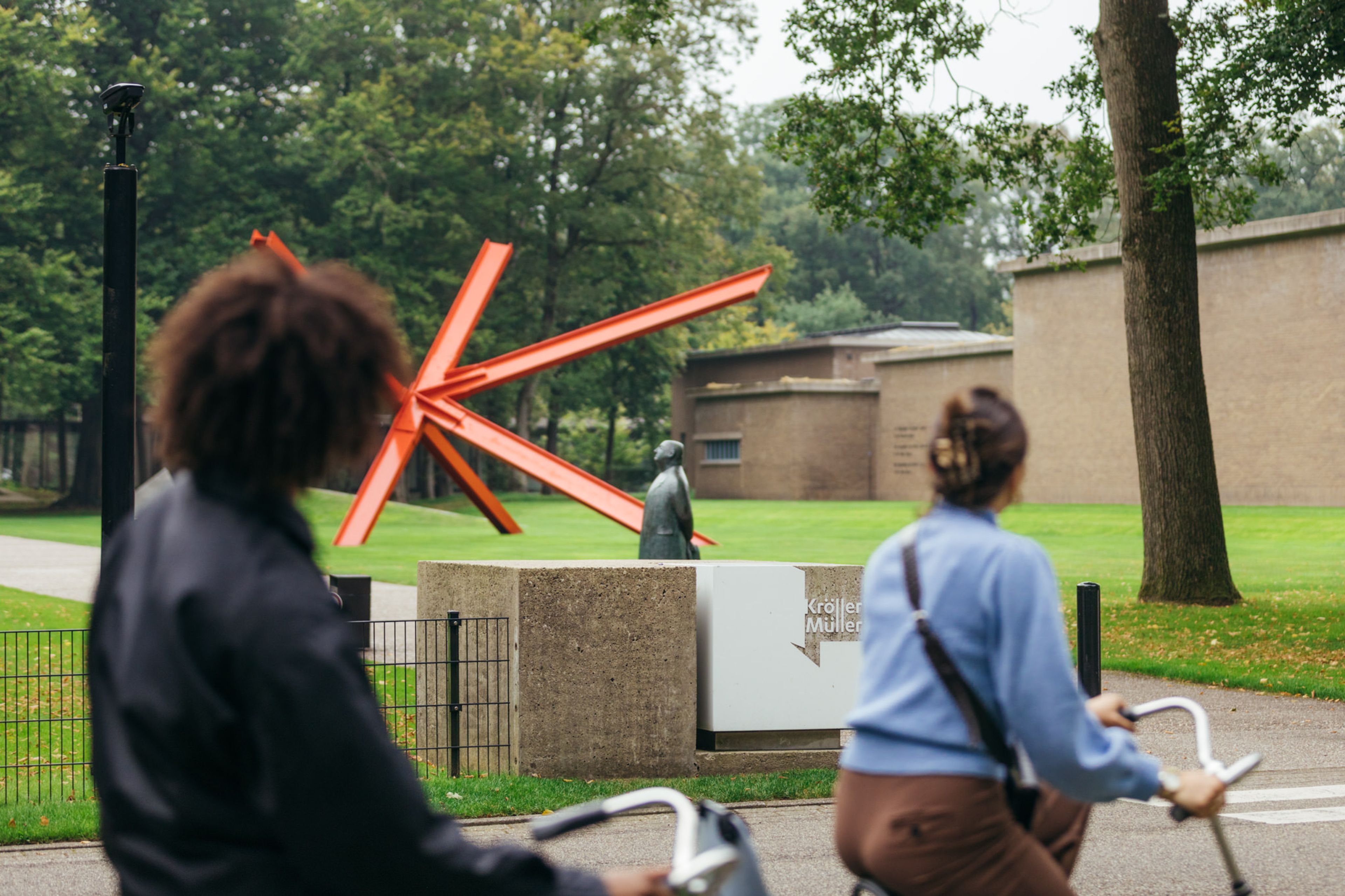 Foto van een buitenkunstwerk bestaande uit grote rode stalen in een parkachtige omgeving. Op de voorgrond twee personen op witte fietsen