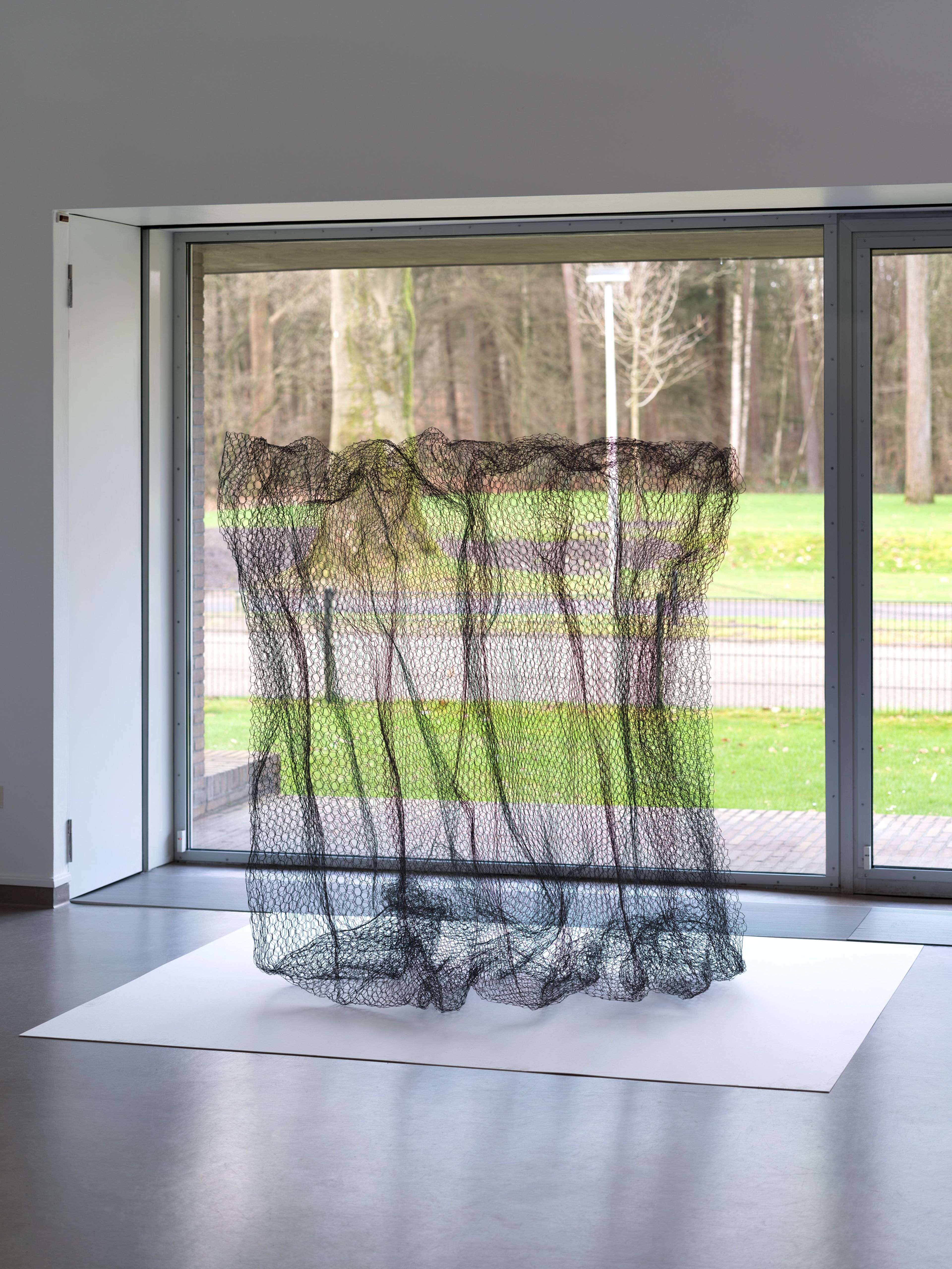 Alan Saret, _Zinc Cloud_, 1967, 1990 Foto van een transparante, golvende draadsculptuur geplaatst op een witte ondergrond in een ruimte met grote ramen en uitzicht op een bosrijke omgeving.