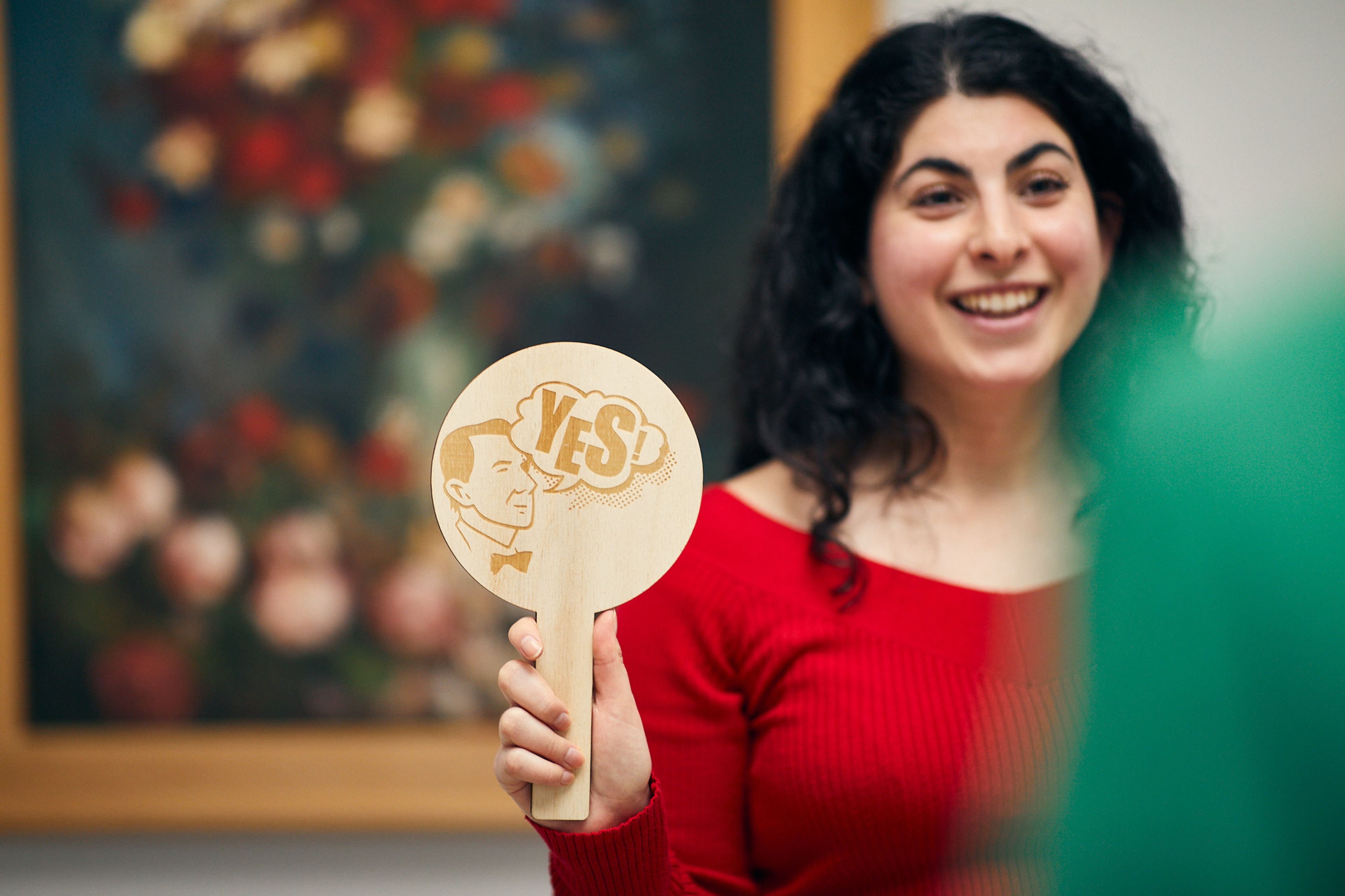 persoon in rode trui die houten bordje vasthoudt met tekst "YES" in een museum- of galerijomgeving. Bordje en persoon centraal, achtergrond met onscherp bloemenschilderij.