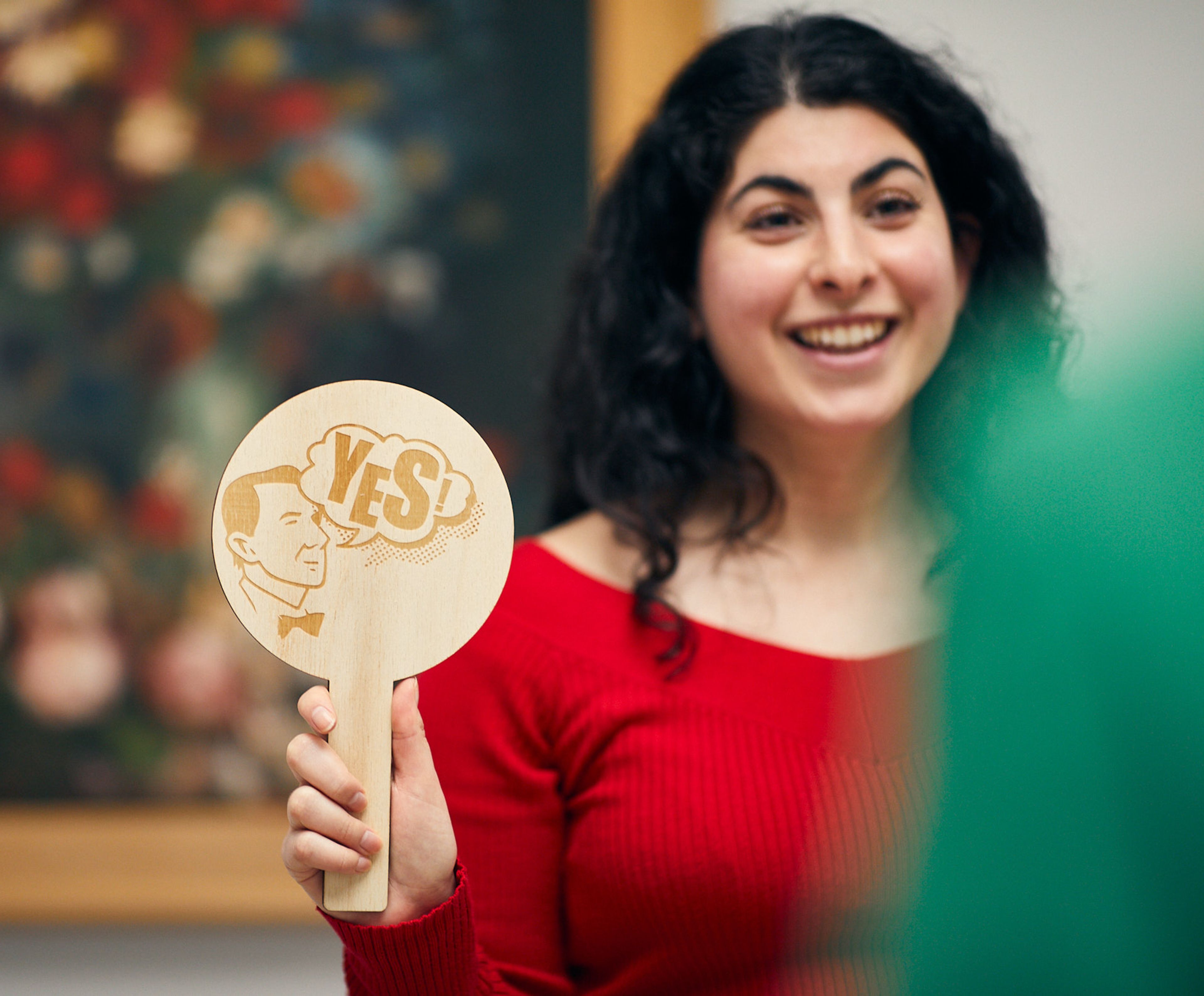 person in red sweater holding wooden sign with text "YES" in a museum or gallery setting. Sign and person central, background with blurred flower painting.