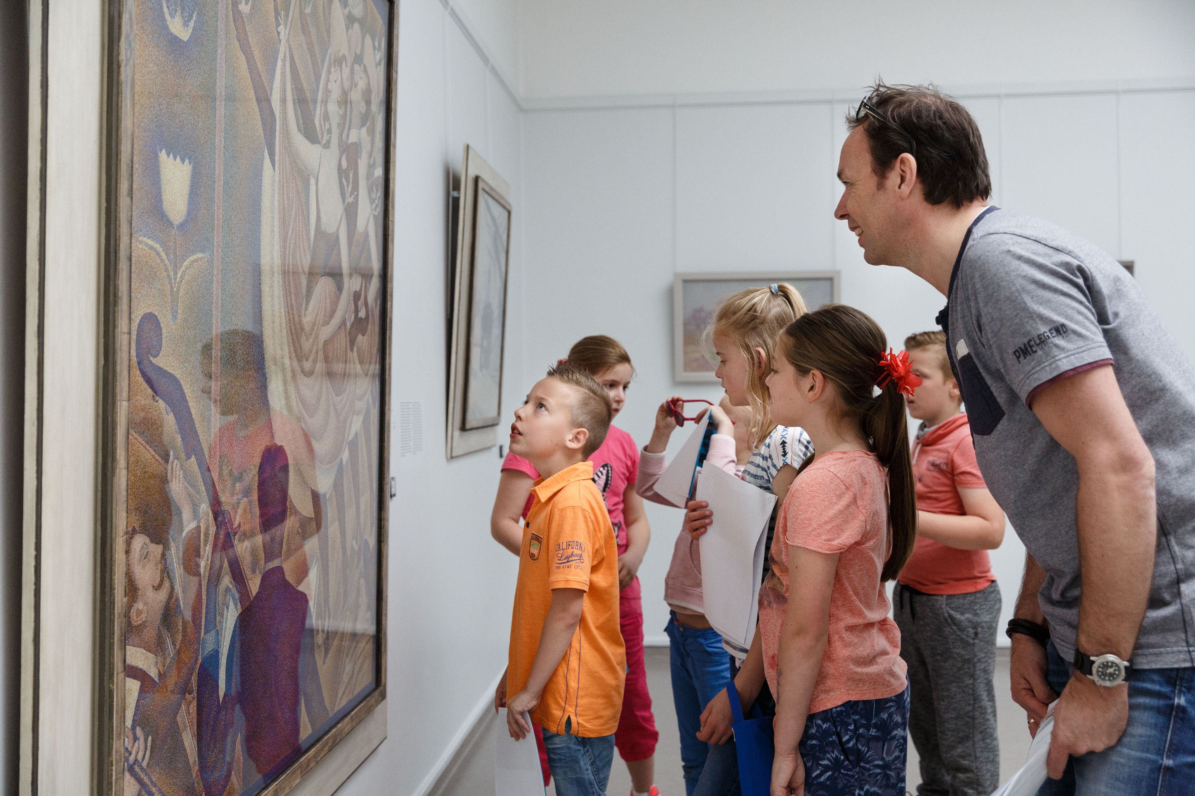 Groep kinderen en een volwassene die aandachtig naar een groot schilderij in een museum kijken. Kinderen dragen kleurrijke kleding en houden tekenmateriaal vast, wat wijst op een educatieve activiteit gericht op kunstbeleving en leren.