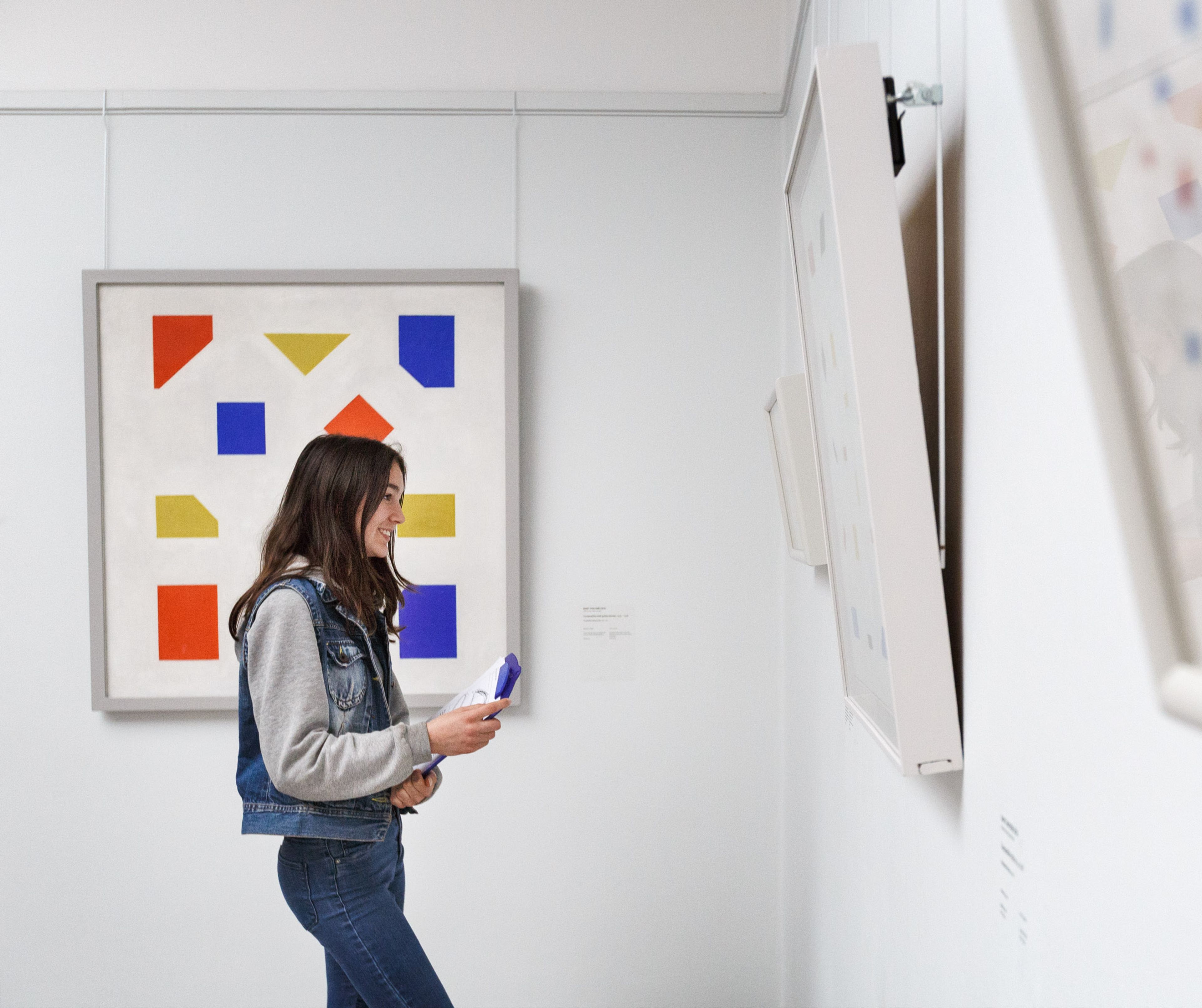 Een persoon die een kunstwerk met kleurrijke geometrische vormen in een galerie bekijkt. Kunstwerk bevat rode, gele en blauwe rechthoeken en driehoeken op een witte achtergrond, met andere kunstwerken zichtbaar aan de muur.
