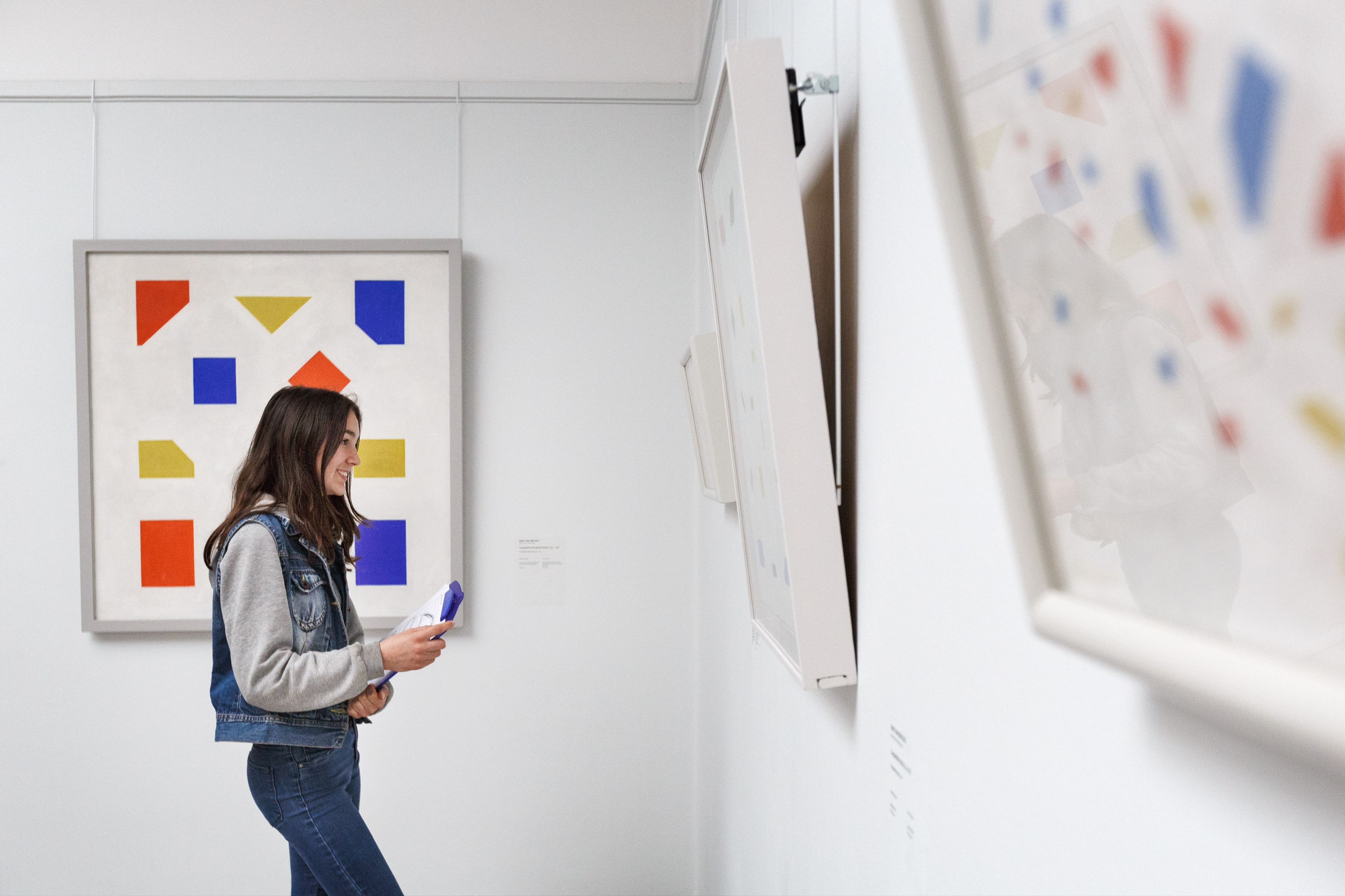 Een persoon die een kunstwerk met kleurrijke geometrische vormen in een galerie bekijkt. Kunstwerk bevat rode, gele en blauwe rechthoeken en driehoeken op een witte achtergrond, met andere kunstwerken zichtbaar aan de muur.
