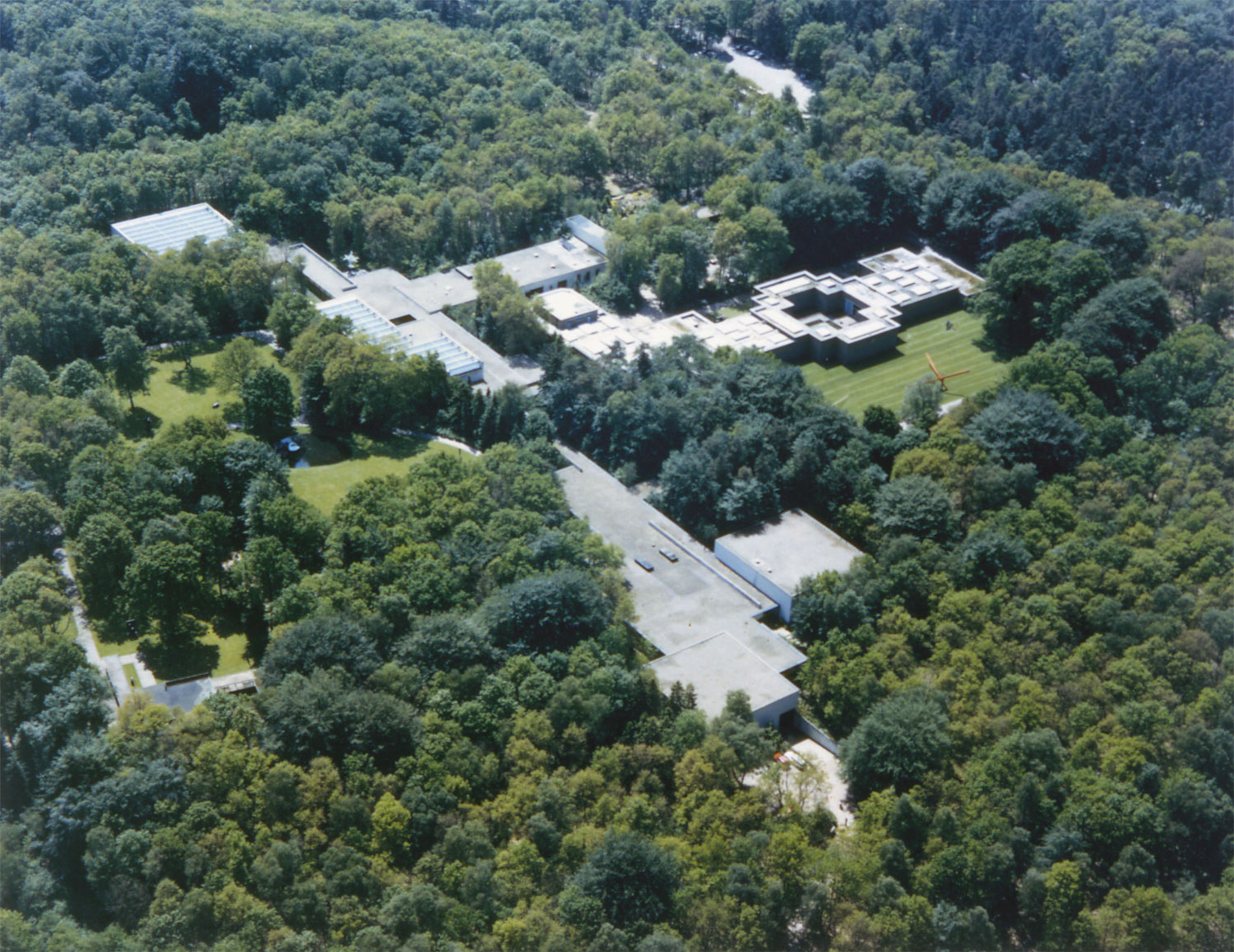 Luchtfoto van het complex van museumgebouwen tussen dicht bos en groen. Structuur met meerdere verbonden delen met platte daken en een opvallend vierkant met binnenplaats.