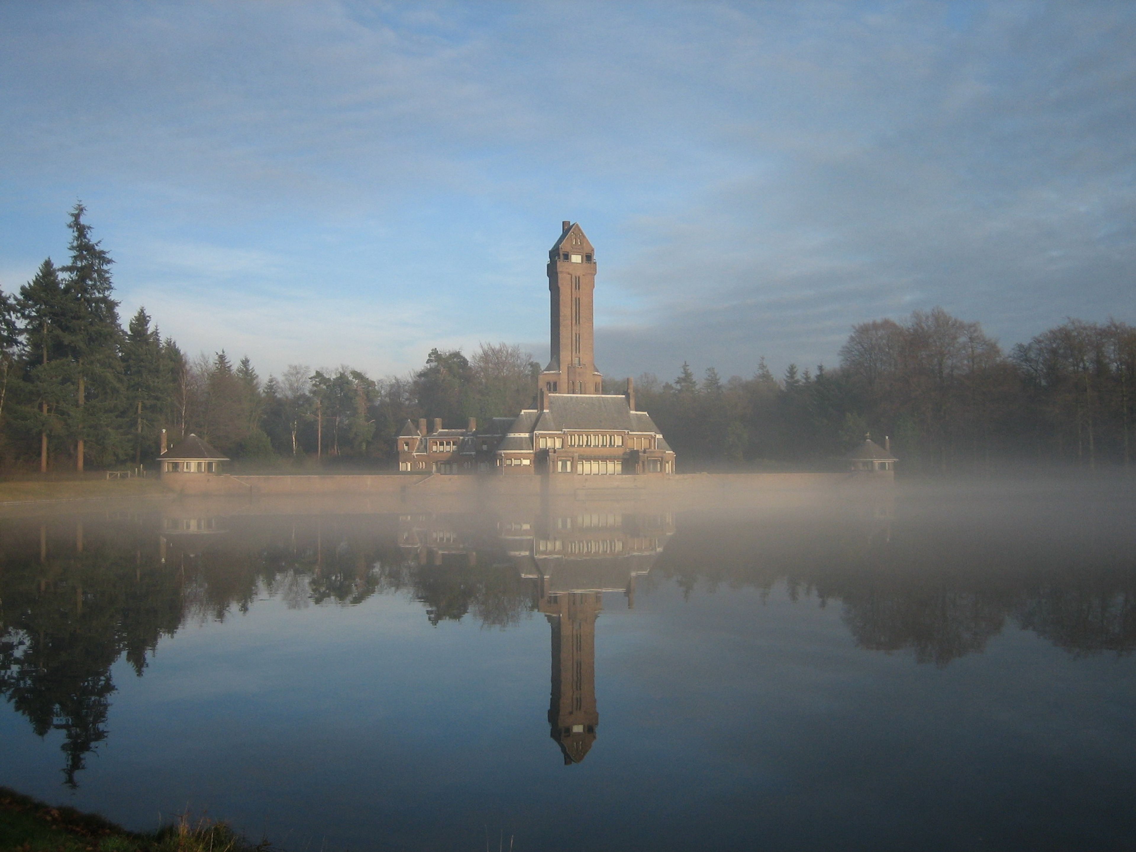 Foto van Jachthuis Sint Hubertus, een toren met omliggende gebouwen, weerspiegeld in een kalm meer met lichte mist erboven. Blauwe lucht en bomen vormen achtergrond