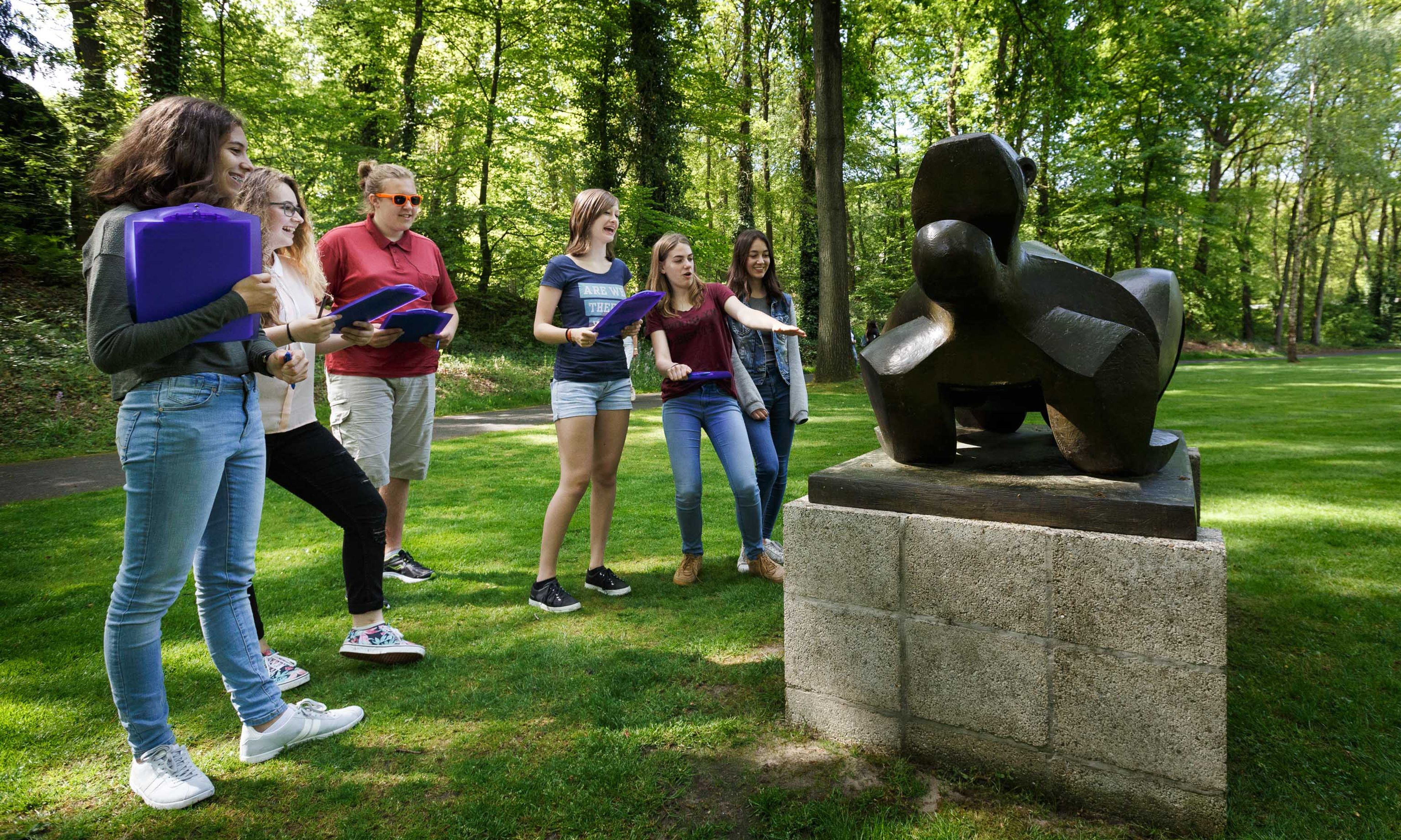 Sculpture garden secondary education Group of six young people looking at and discussing abstract bronze sculpture on concrete pedestal in park with green trees and grass. Young people wear casual clothing and hold folders, indicating educational or cultural activity outside.