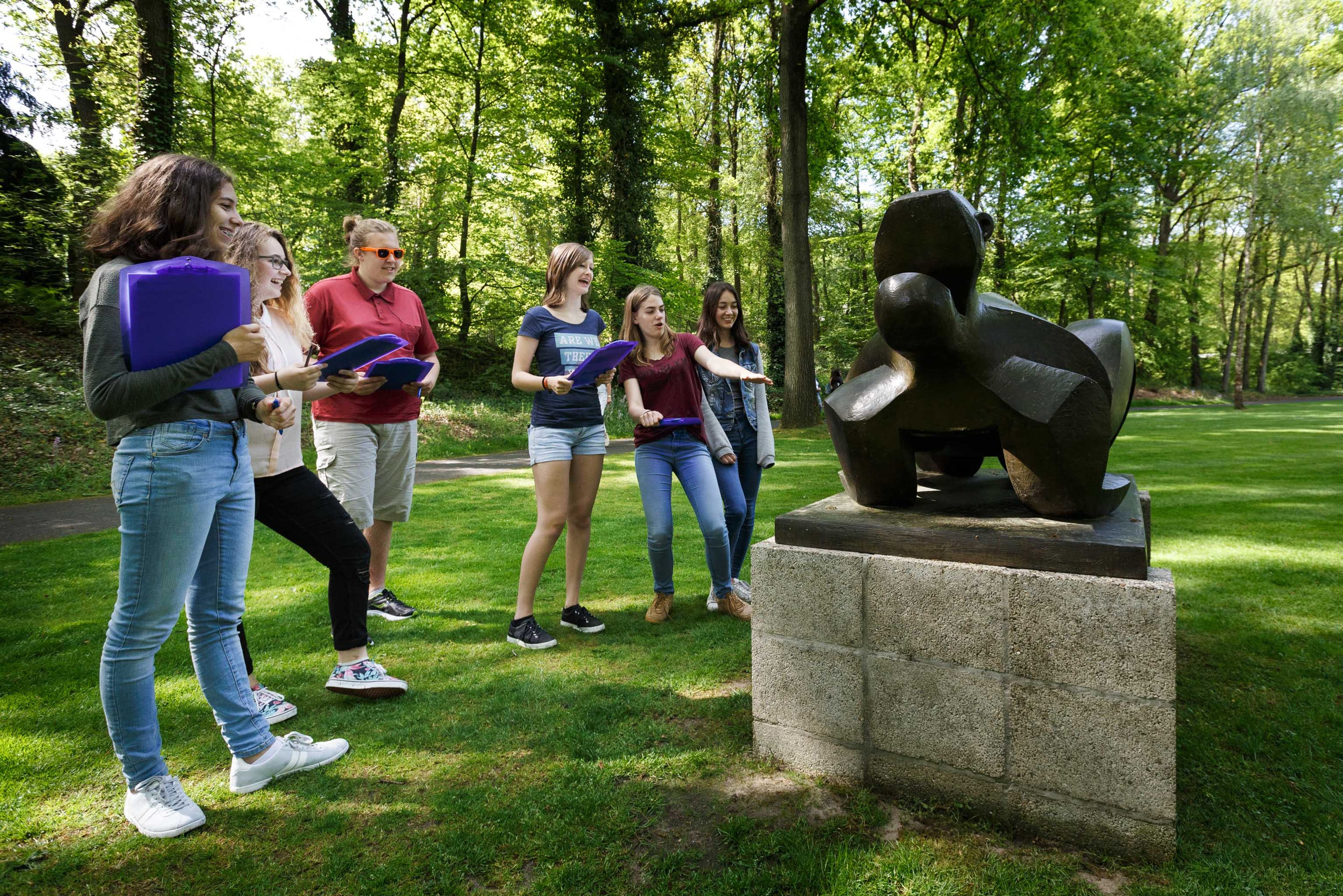 Beeldentuin voortgezet onderwijs Groep van zes jongeren bekijkt en bespreekt abstracte bronzen beeldhouwwerk op betonnen sokkel in park met groene bomen en gras. Jongeren dragen casual kleding en houden mappen vast, wat wijst op educatieve of culturele activiteit buiten.
