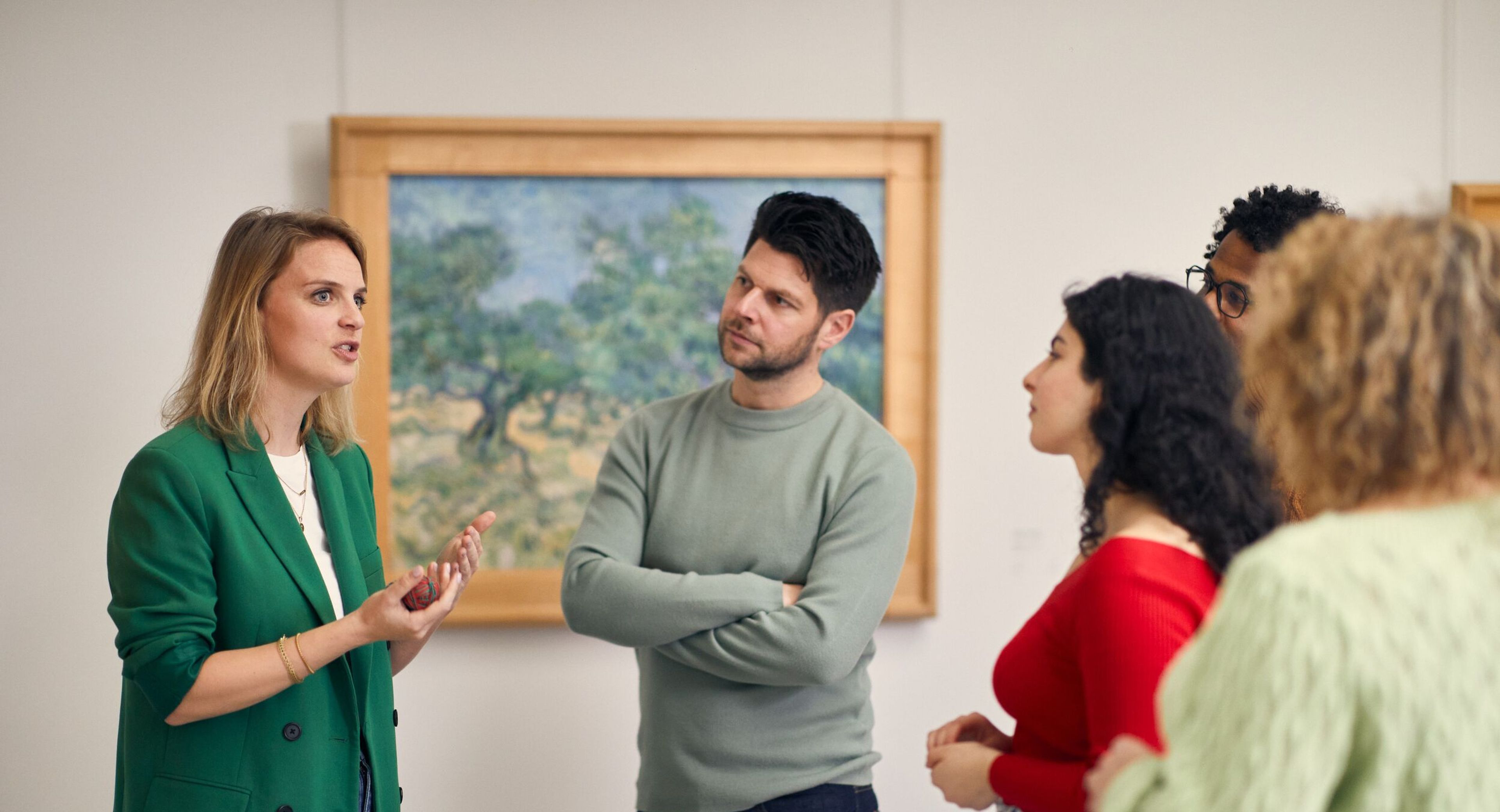 A tour guide talks to a group of people in the Van Gogh gallery. In the background is a painting by Van Gogh depicting olive trees in green, blue and yellow.
